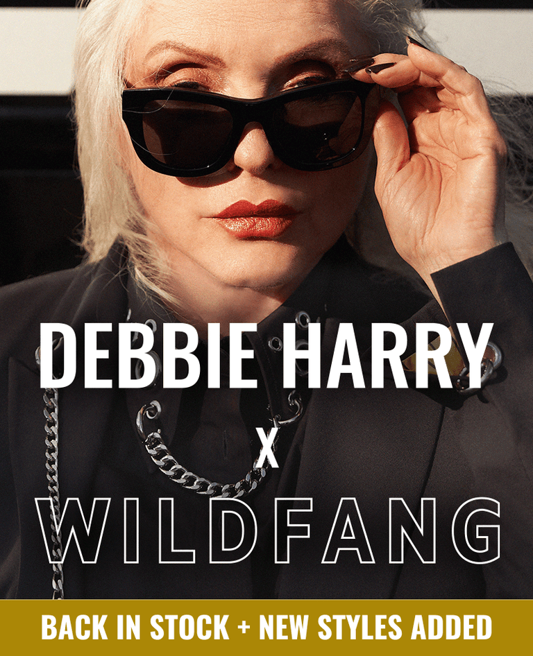 Close up of Debbie Harry looking at camera wearing sunglasses and pieces from the Debbie Harry x Wildfang collection. Text overlay says DEBBIE HARRY X WILDFANG back in stock + new styles added.