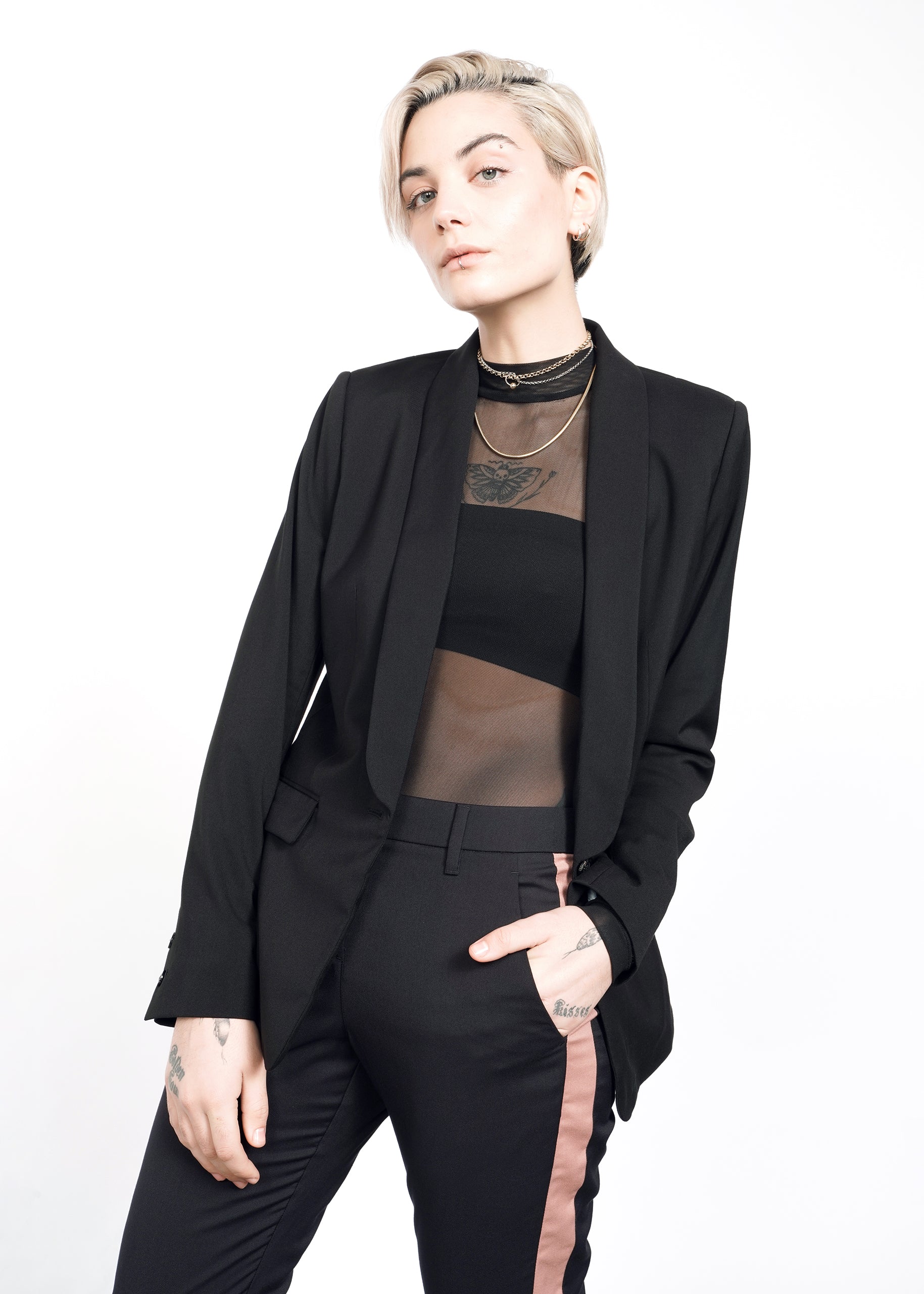 With short blonde hair, they wear The Empower Tux Blazer, a sheer black top, and black pants with pink stripes. Tattoos on their hand and wrist are complemented by various necklaces, all set against a white backdrop.