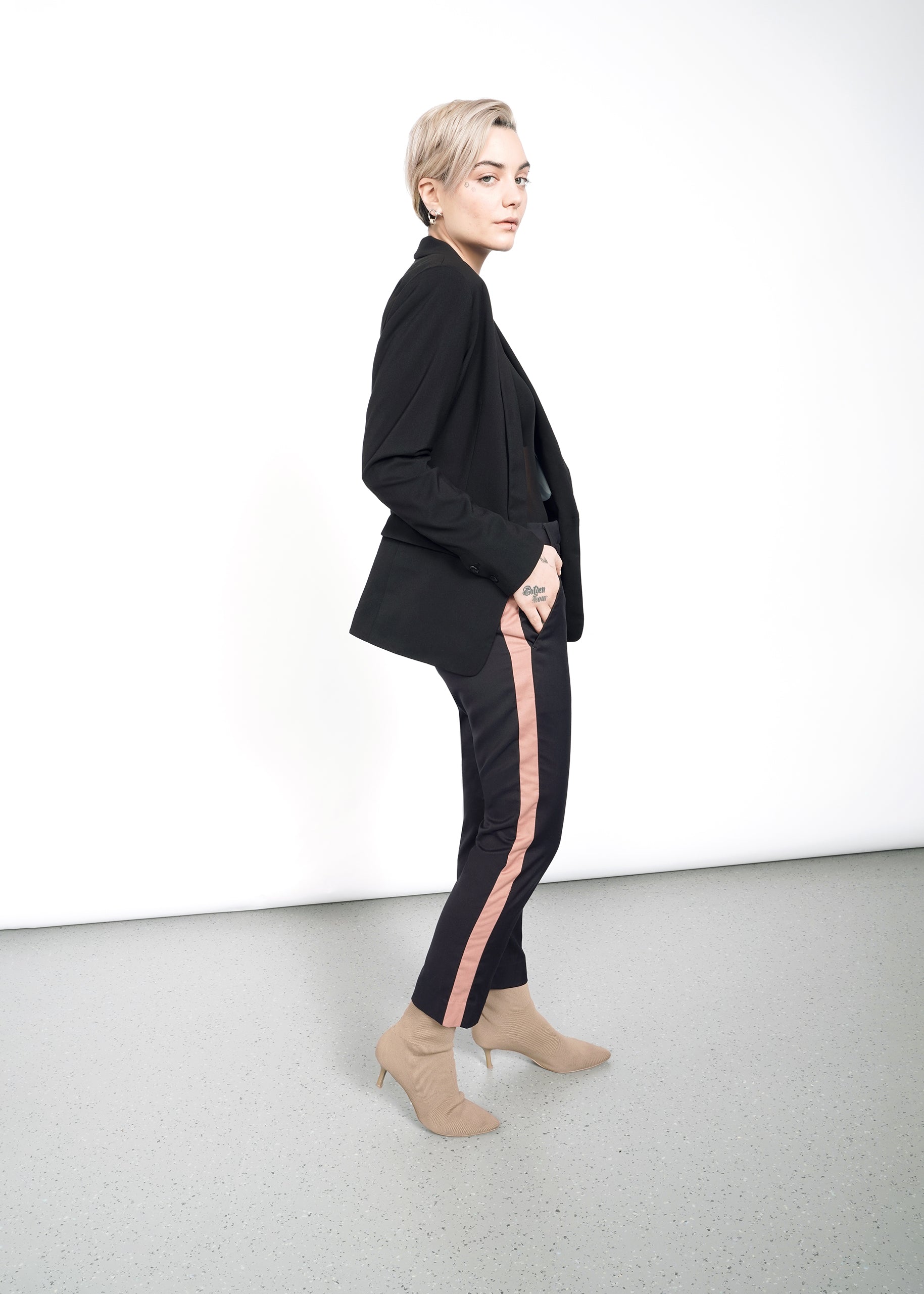 A person with short hair wears The Empower Tux Blazer in black, featuring a pink stripe detail, paired with beige ankle boots. They stand against a white wall, flaunting deep pockets on the timeless blazer and showcasing a hand tattoo.