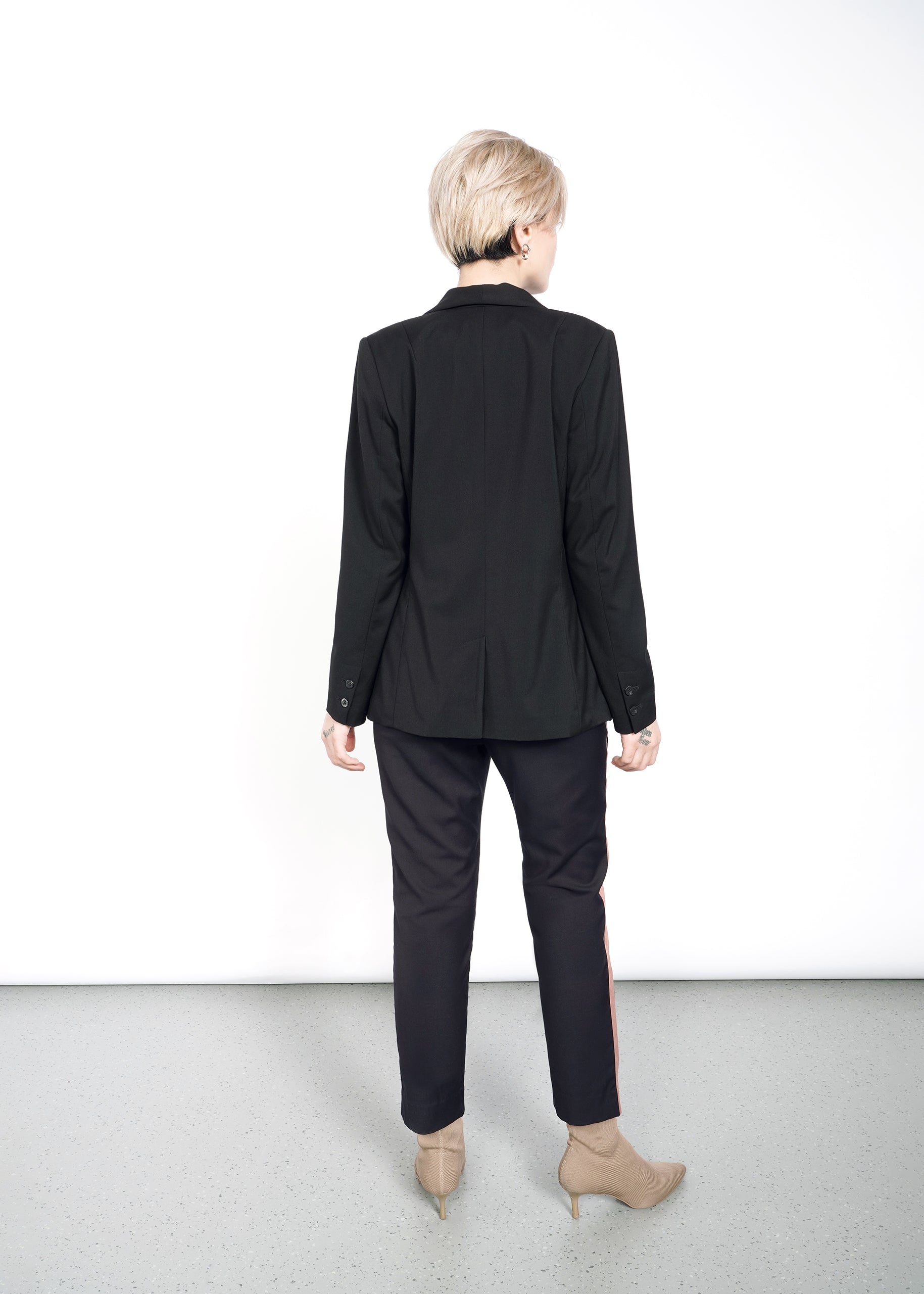 A person with short blonde hair, wearing The Empower Tux Blazer, black pants, and beige ankle boots, stands with their back to the camera. The tailored black jacket exudes timeless elegance as they stand on a gray floor against a white background.