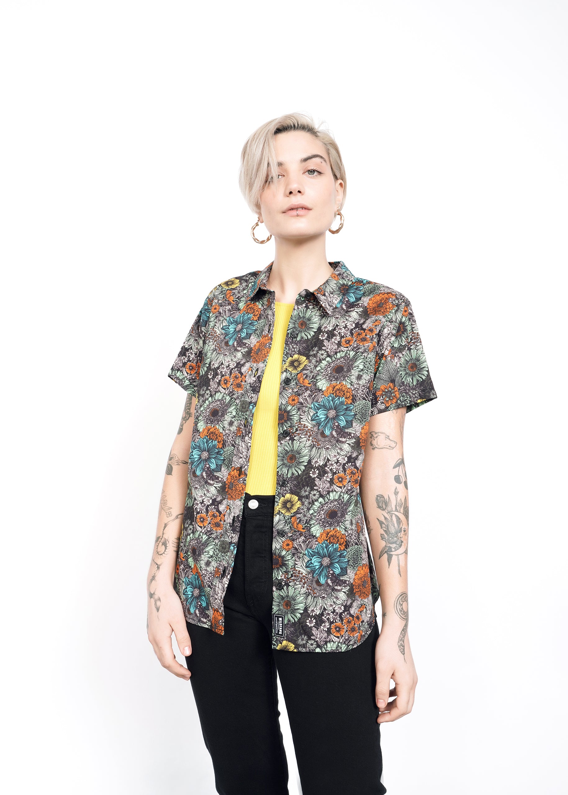 A person with short blonde hair stands confidently against a white background, wearing The Essential Button Up—a tailored-fit, floral-patterned shirt made of 100% cotton—with a yellow top and black pants. They have tattoos on both arms and are wearing hoop earrings.