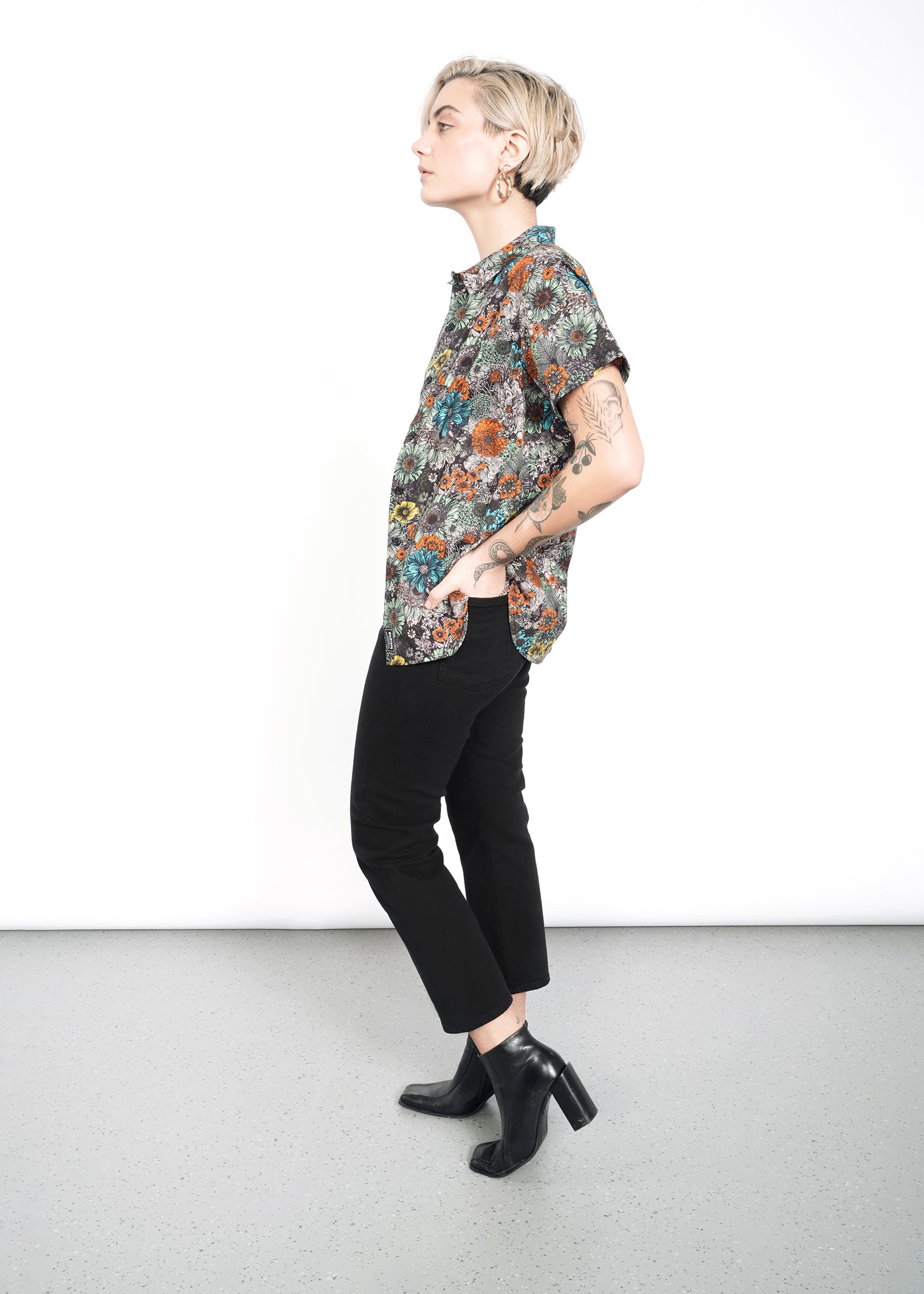 A person with short blonde hair stands in profile against a white background, showcasing The Essential Button Up, a patterned short-sleeve shirt with a classic collar. The 100% cotton fabric complements their black pants and ankle boots, while tattoos add flair to the tailored fit ensemble.