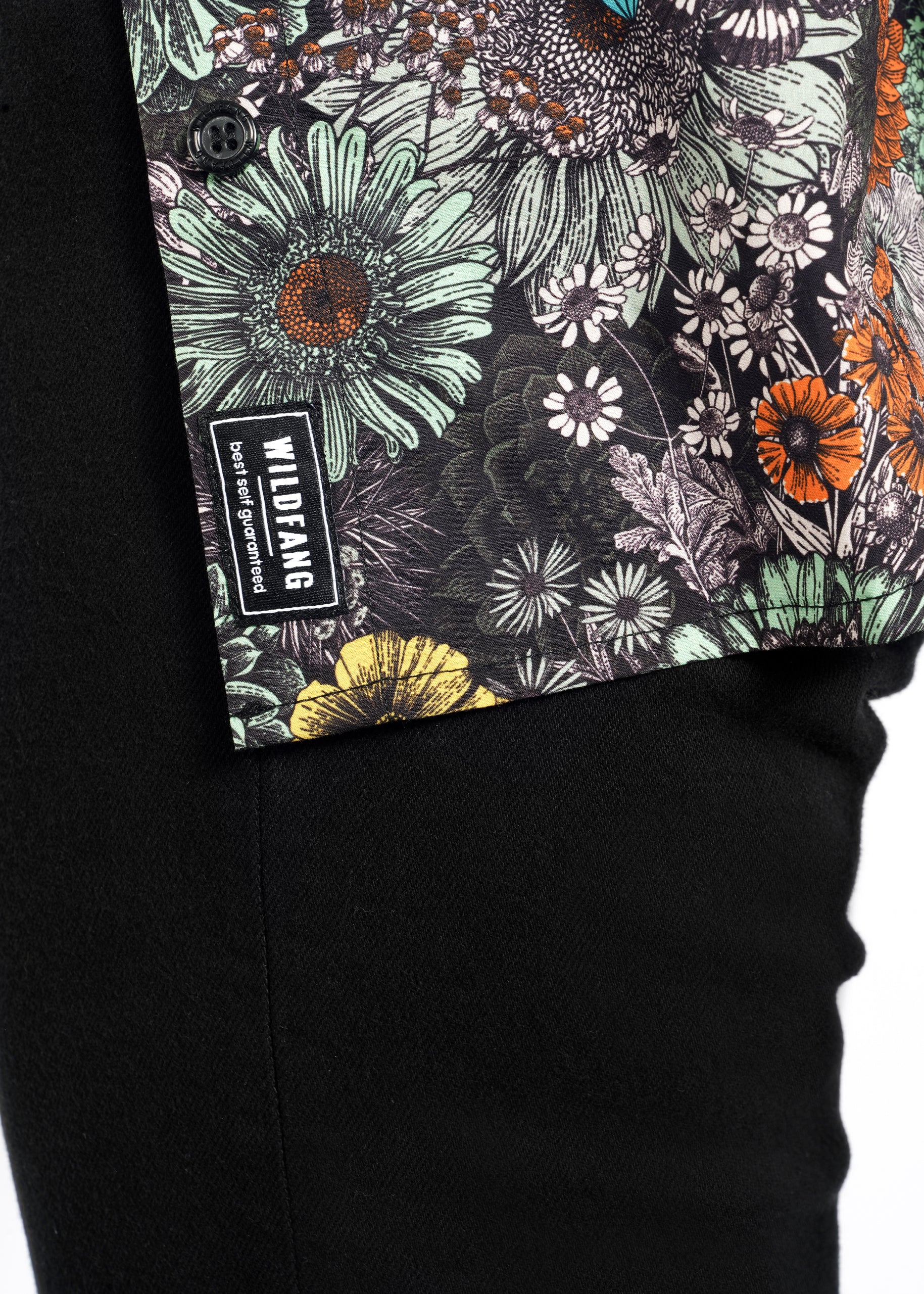 A close-up shows a person in black pants and The Essential Button Up floral shirt featuring green, orange, yellow, and white flowers. The classic collar enhances the tailored fit and the WILDFANG tag stands out. Made from 100% cotton, this outfit offers comfortable style.