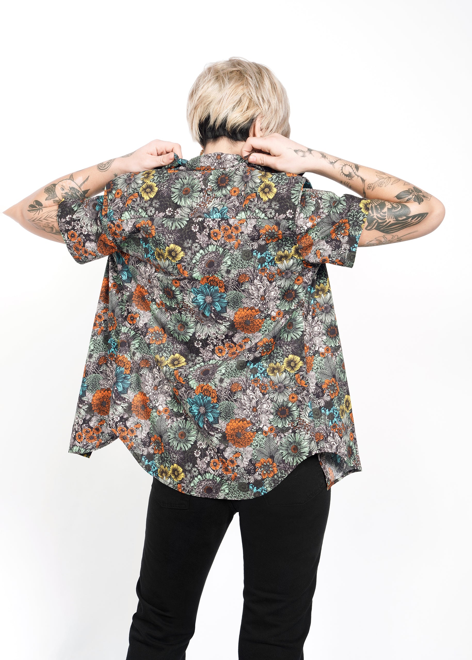 A person with short blonde hair and arm tattoos is wearing The Essential Button Up, a tailored fit, colorful floral-patterned shirt in 100% cotton, paired with black pants. They face away from the camera, lifting their classic collar against a white background.