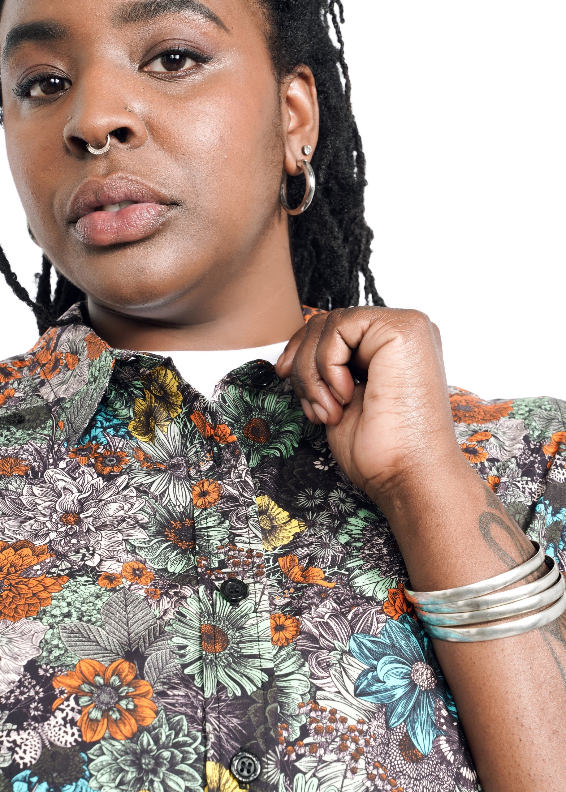 A person with dreadlocks and hoop earrings wears The Essential Button Up, a colorful, floral-patterned 100% cotton shirt. They have a nose ring and hold the classic collar of their tailored fit shirt. A tattoo and silver bangles adorn their arm against a white backdrop.