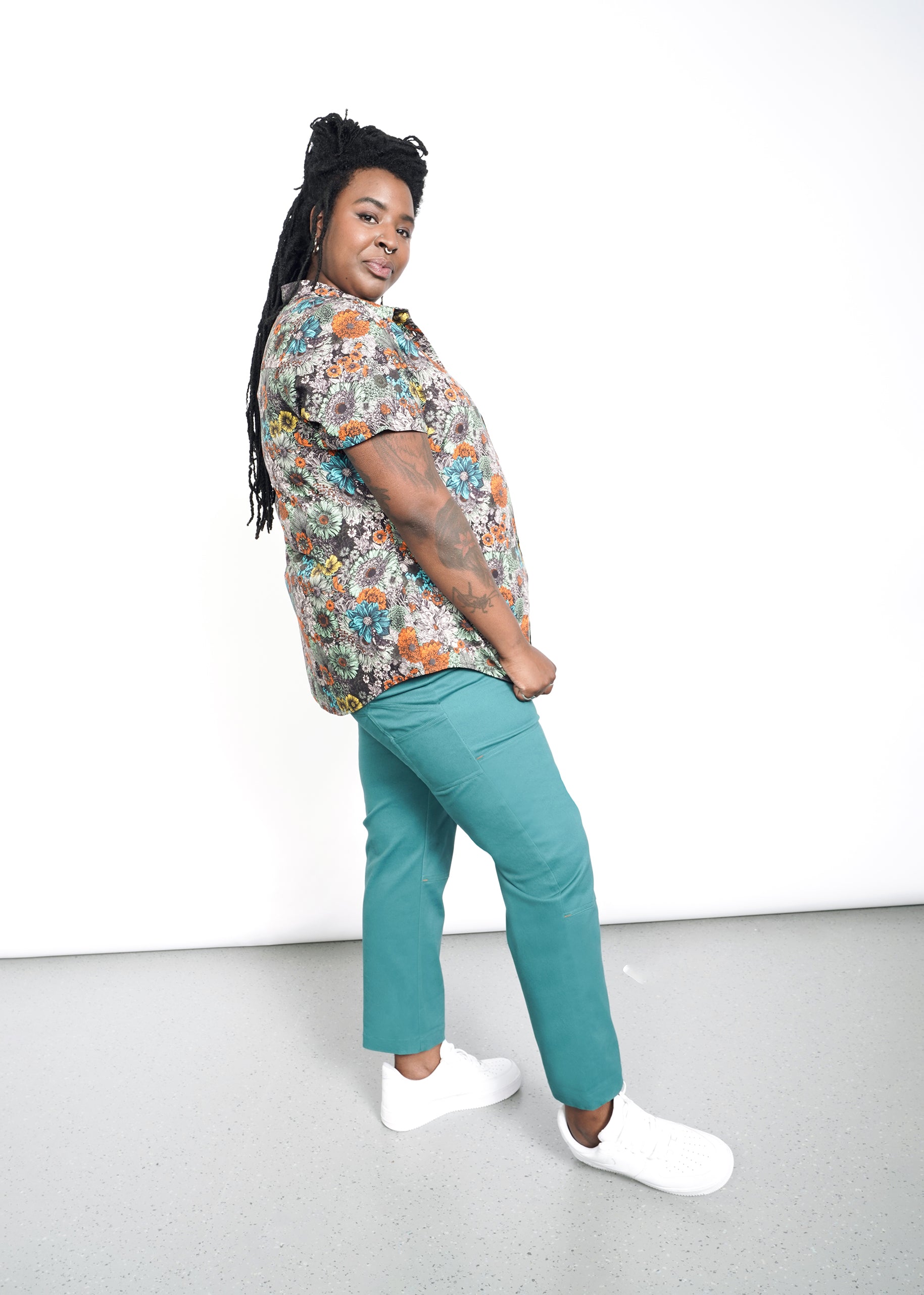 A person with long braided hair stands smiling in a side profile, wearing The Essential Button Up featuring a classic collar. Paired with teal pants and white sneakers, the tailored fit enhances their elegance against the minimalist white background.