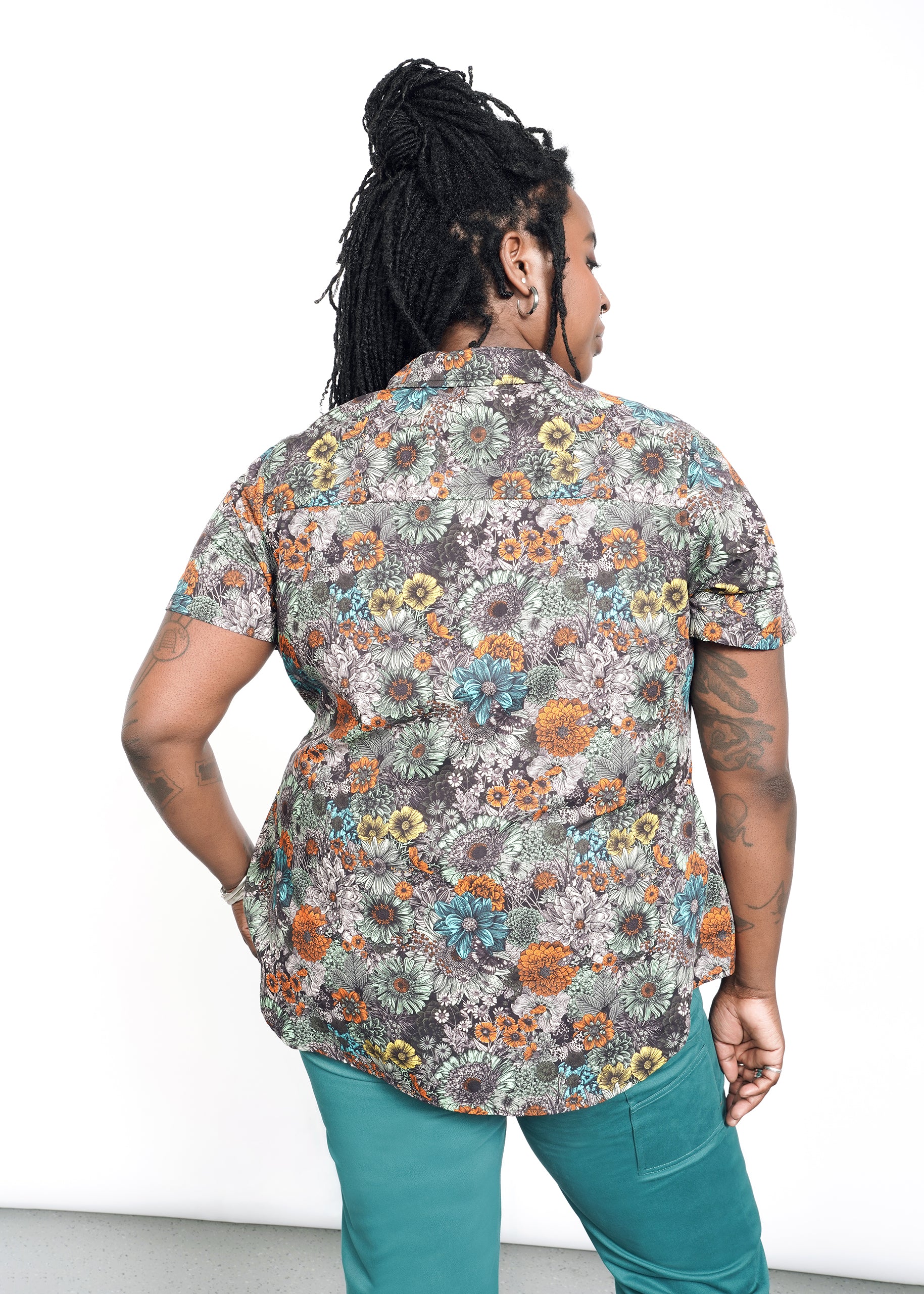 A person with long braided hair and tattoos on their left arm is facing away, wearing The Essential Button Up, a short-sleeved floral cotton shirt with a classic collar and tailored fit, paired with teal pants. Their right hand rests on their hip against a plain white background.