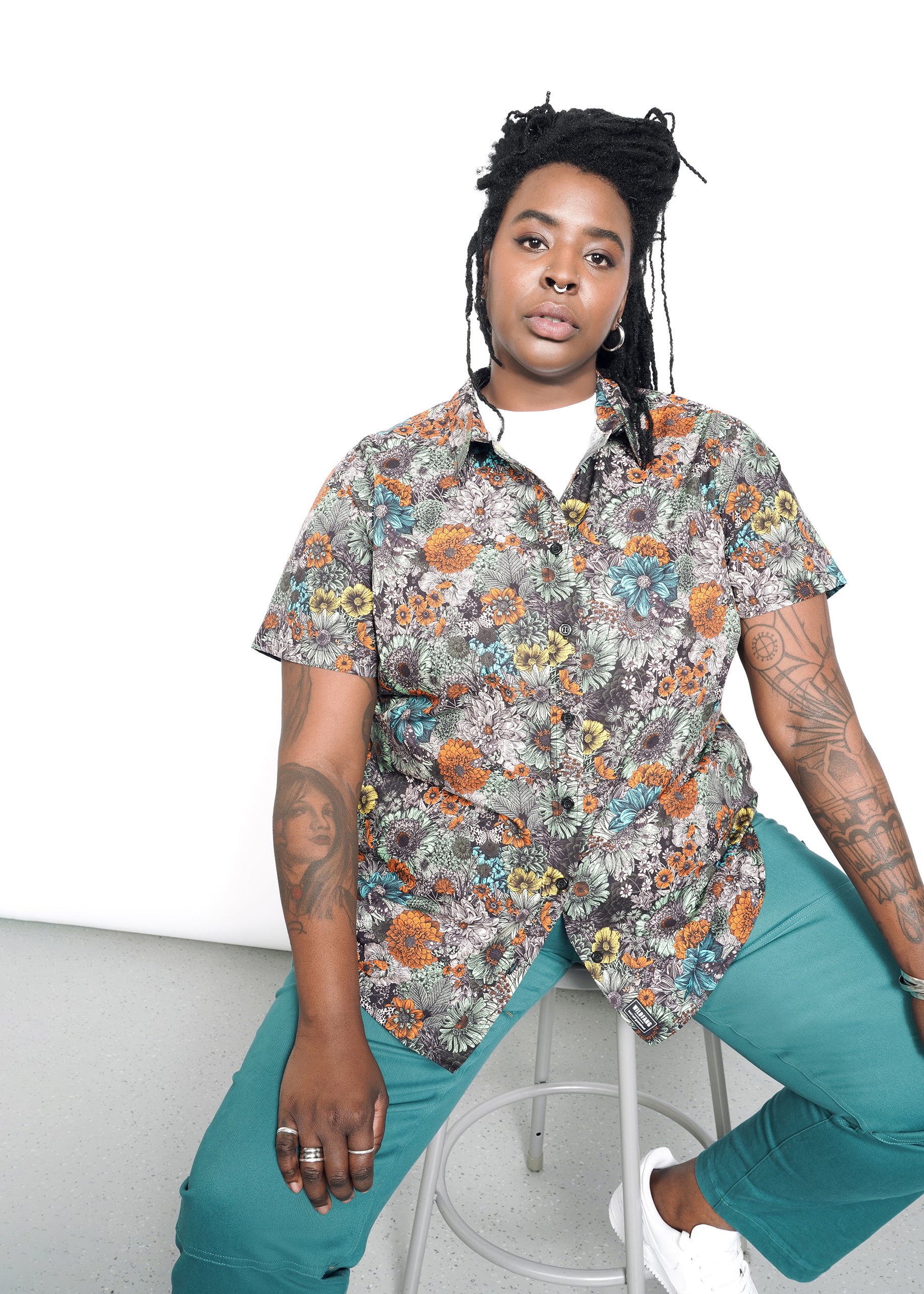 A long-haired person, dressed in The Essential Button Up—a tailored fit, floral-patterned short-sleeve 100% cotton shirt—and teal pants, sits on a stool. White sneakers are visible. A plain white background accentuates colorful tattoos and the shirts classic collar.