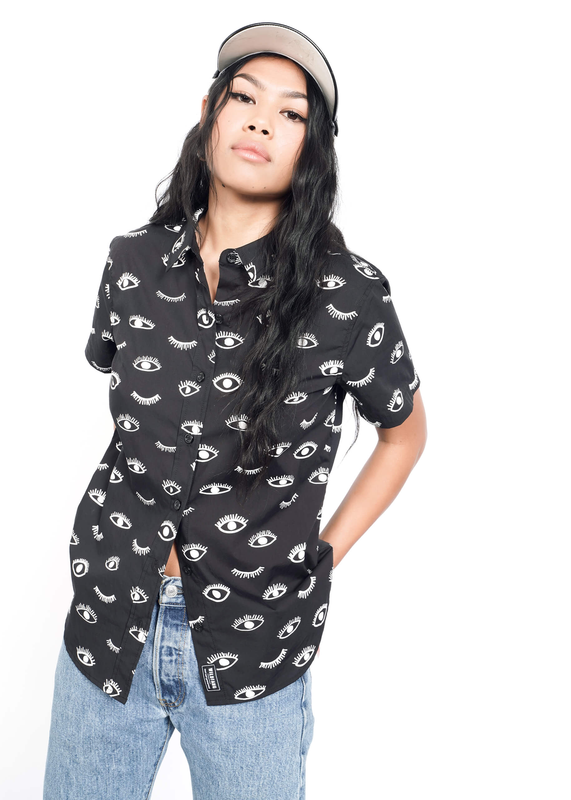 A person in a black Wildfang The Essential Button Up shirt with eye designs, light blue jeans, beige cap, and long hair stands against a plain white background with hands in pockets.