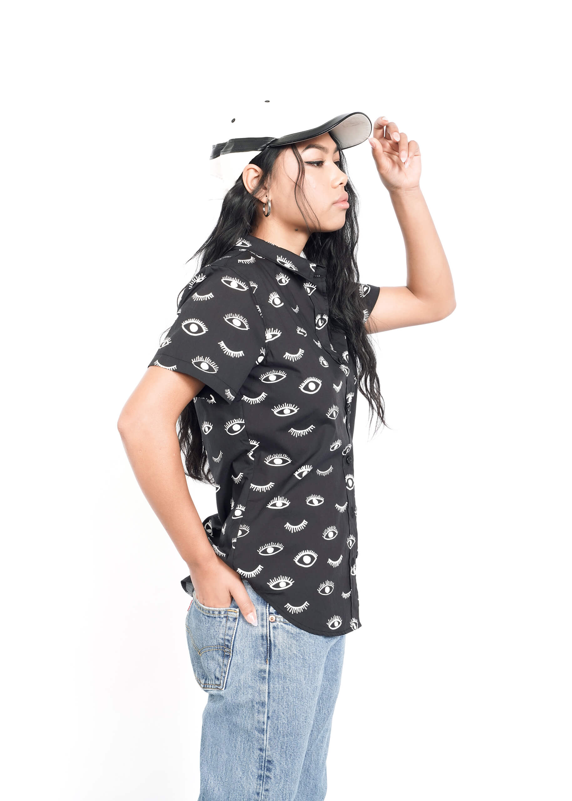 A person with long hair stands in profile against a white background, wearing The Essential Button Up shirt from Wildfang with eye patterns, blue jeans, and a cap. One hand is in their pocket while the other adjusts the cap.