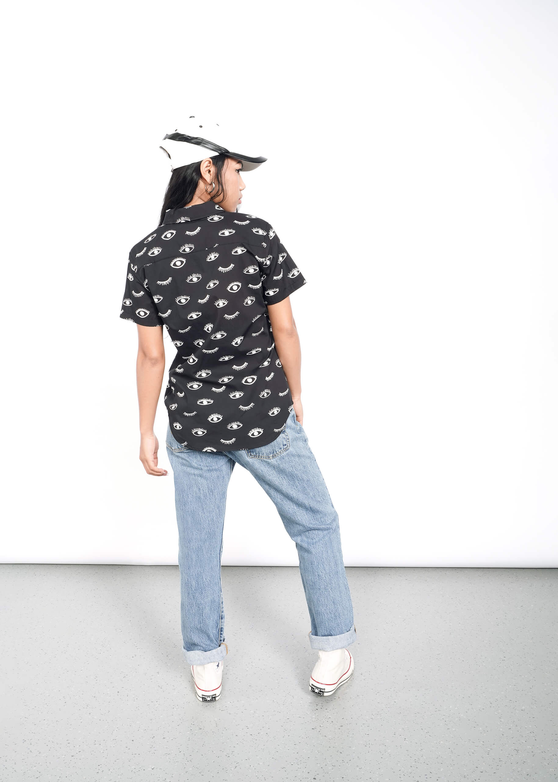 Sporting a black Wildfang The Essential Button Up with an eye pattern, tailored fit light blue jeans, and white sneakers, someone stands with their back to the camera against a plain white background wearing a stylish black and white cap.