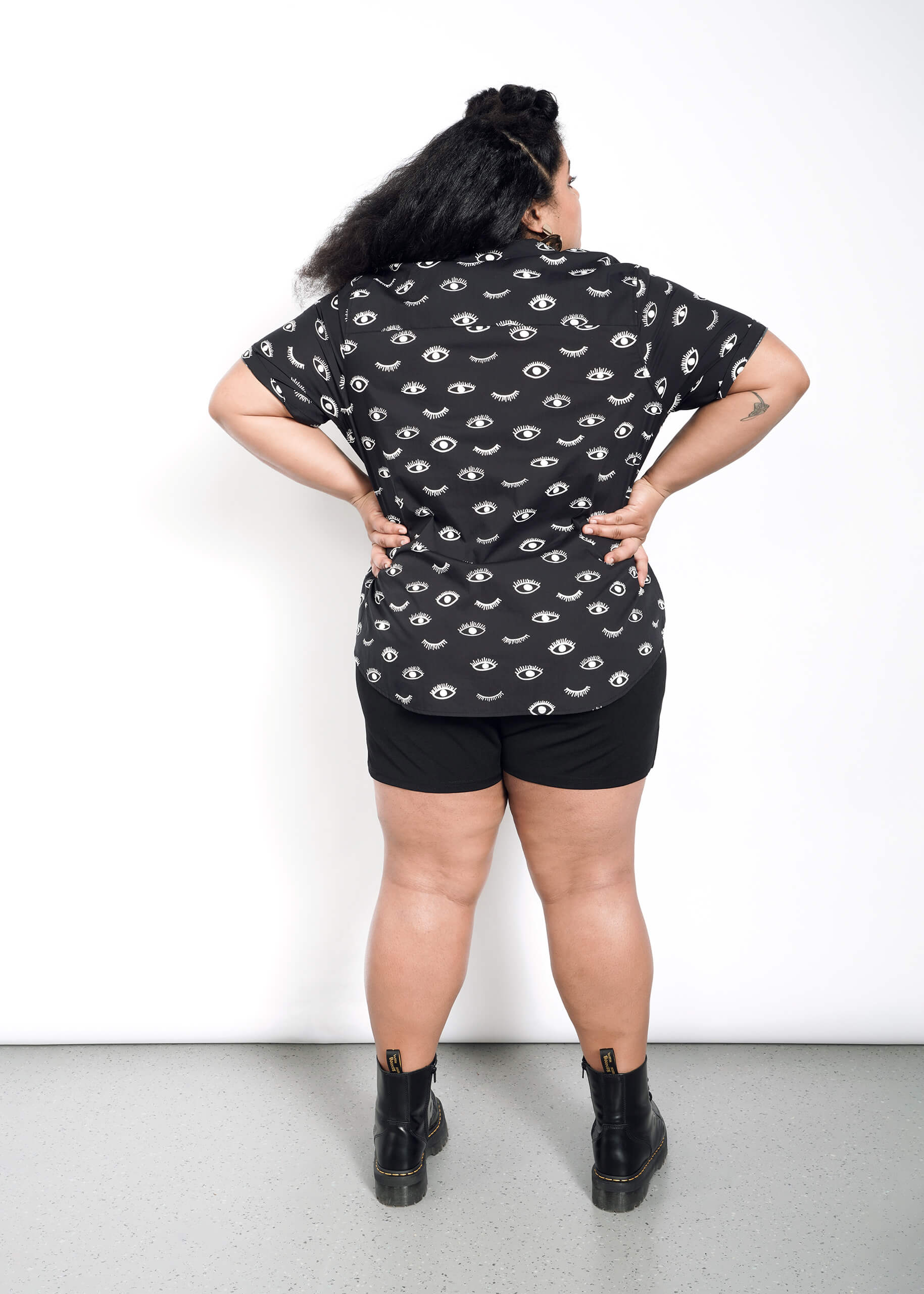 A person with long dark hair stands against a white wall, facing away. They wear a tailored fit Wildfang The Essential Button Up shirt decorated with eye patterns, black shorts, and black boots. One hand rests on their hip.