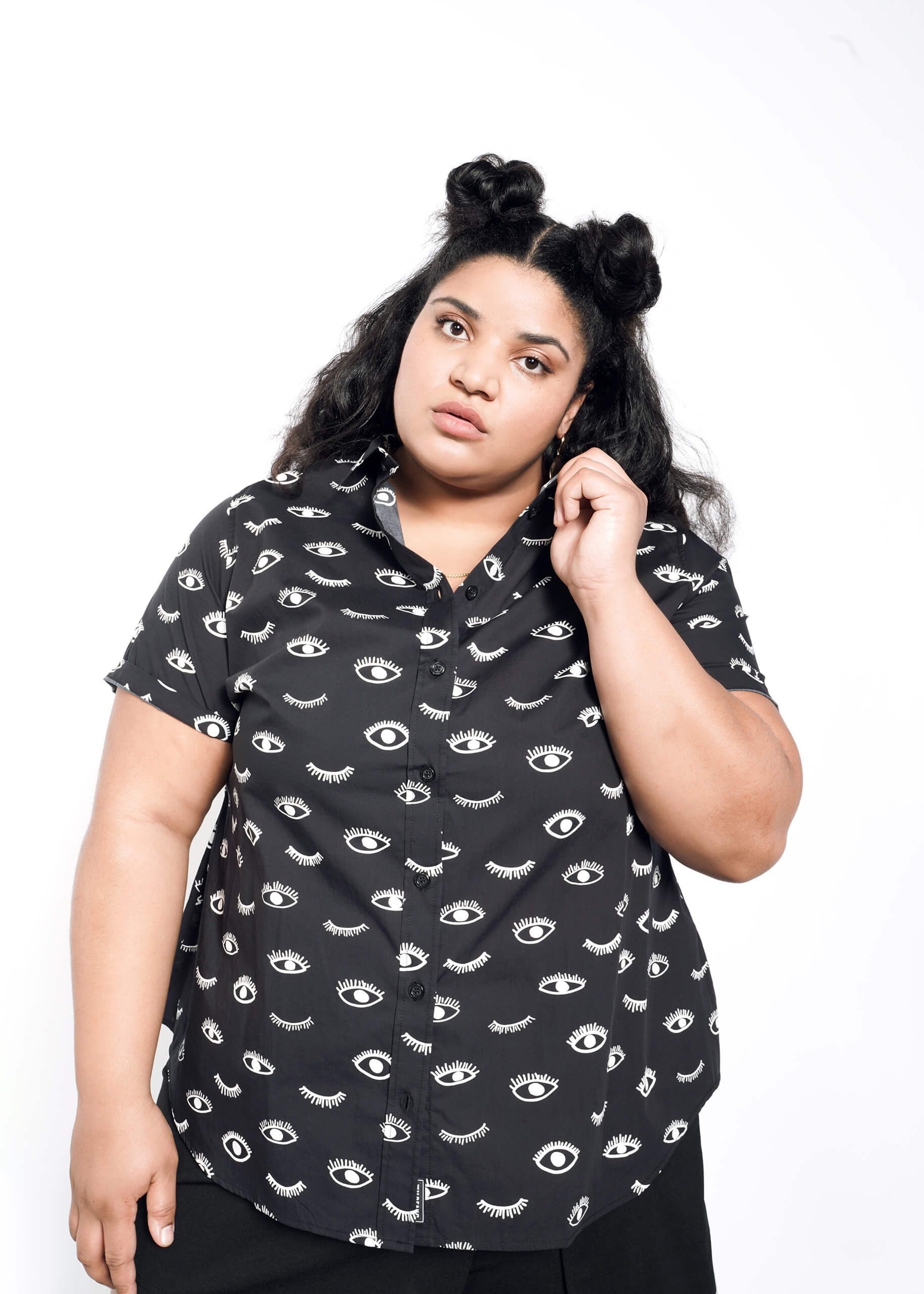 A person with long, dark hair styled in two buns wears The Essential Button Up from Wildfang, featuring a classic eye pattern. They are facing the camera, holding the collar of their tailored-fit black shirt against a white background.