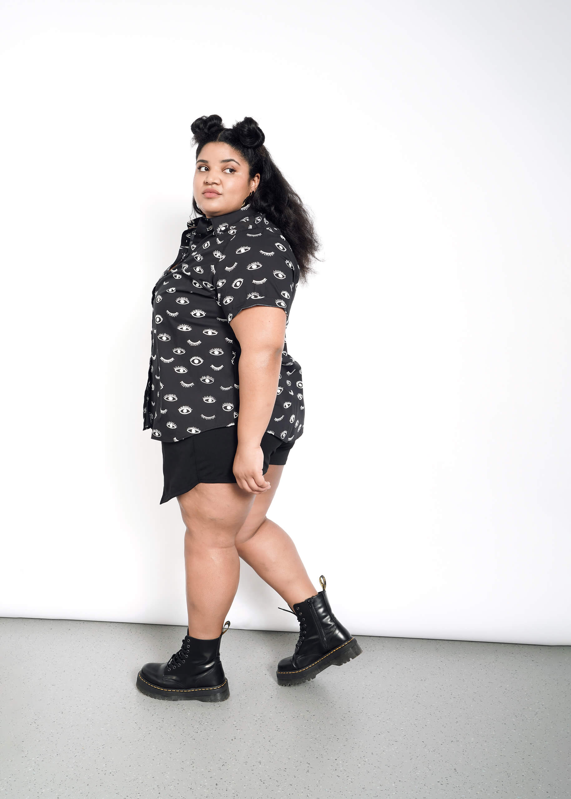 A long-haired individual with styled buns wears a tailored fit Wildfangs The Essential Button Up shirt adorned with eye patterns, paired with black shorts and boots, standing against a white background while looking to the side.