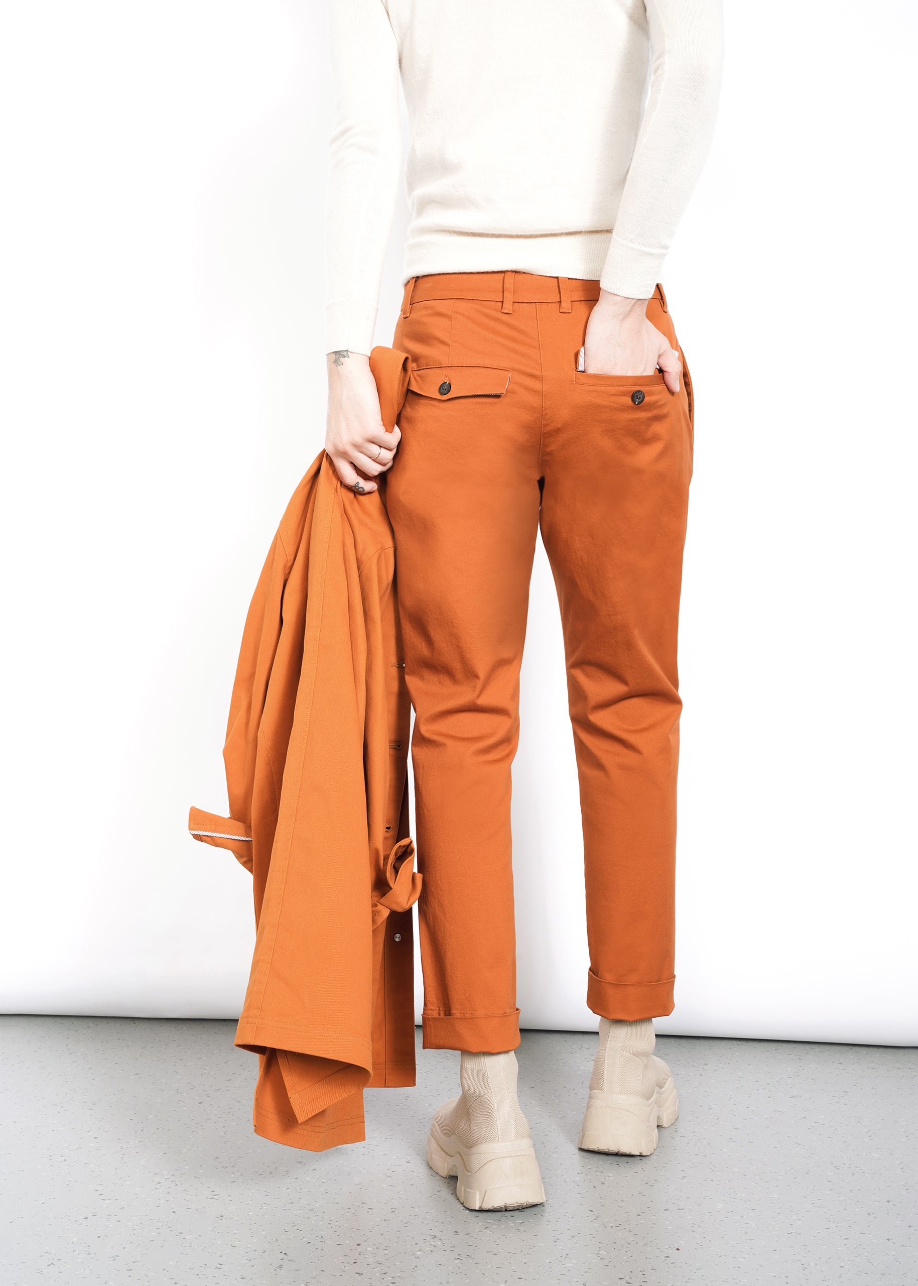 A person wearing The Essential Trouser in a relaxed-fit orange style, paired with a white sweater. Theyre holding a matching orange jacket in their left hand and are wearing beige shoes, all against a plain white background.