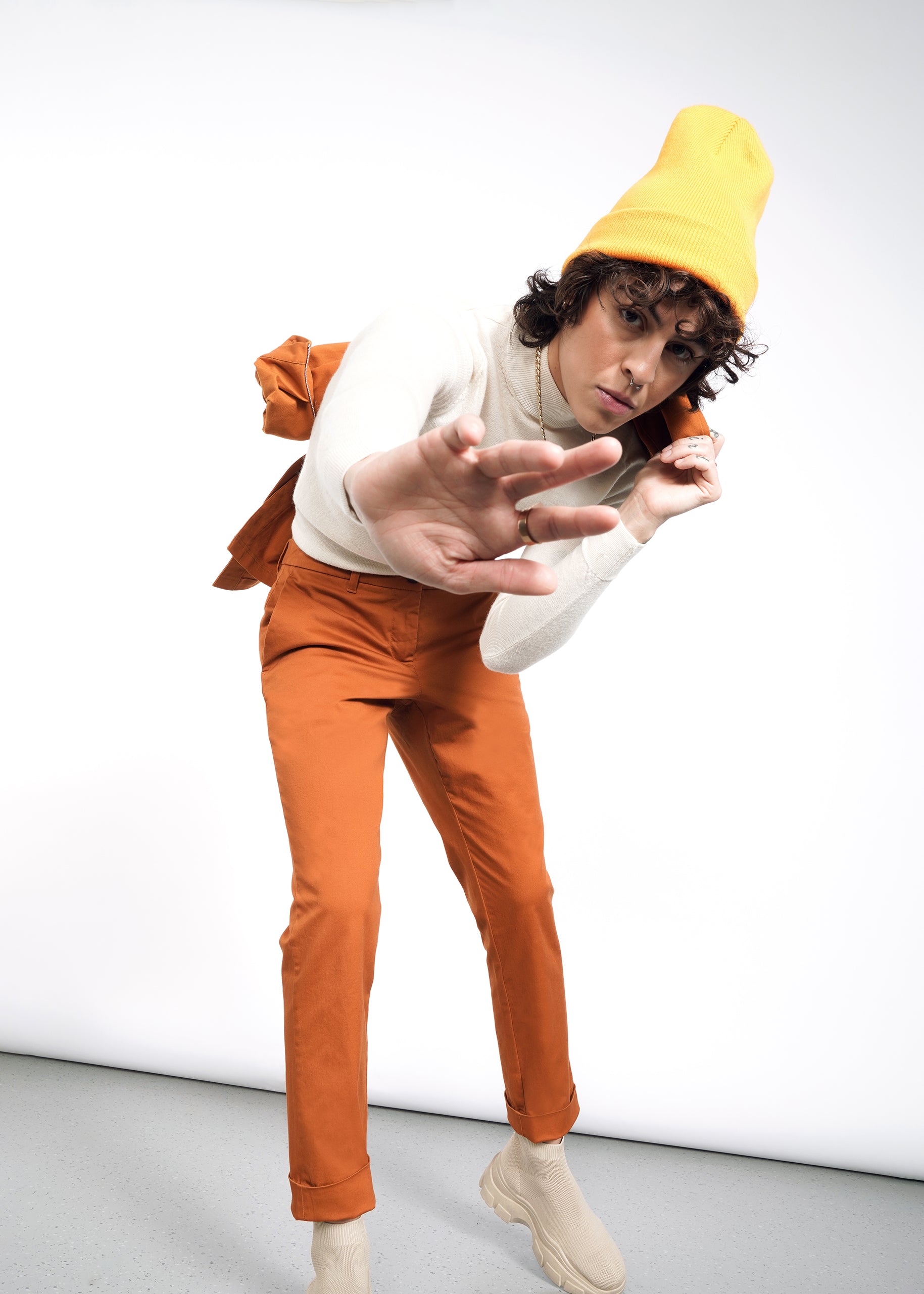 Wearing a mustard-yellow beanie, orange The Essential Trouser, a white sweater, and beige shoes, a person playfully leans forward and reaches toward the camera against a plain white background.