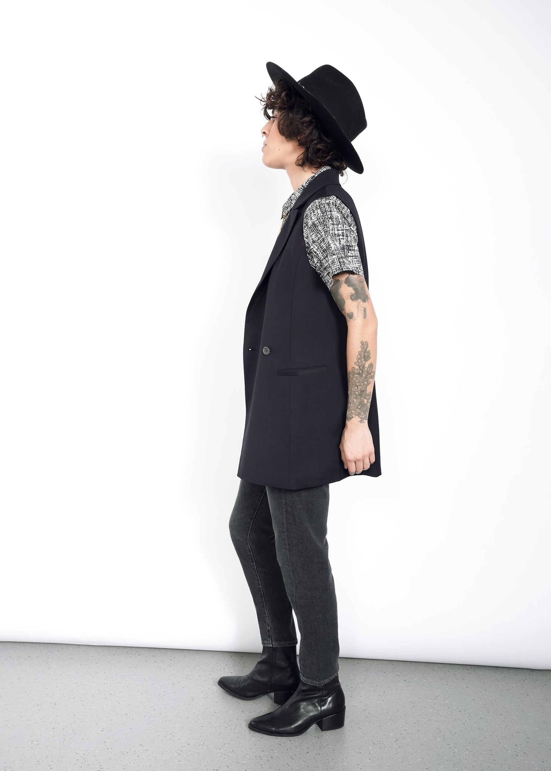 With curly hair and tattoos, a person in profile against a white background wears The Empower Double Breasted Vest in an oversized fit, paired with a patterned shirt, black jeans, boots, and a black hat while gazing upwards.