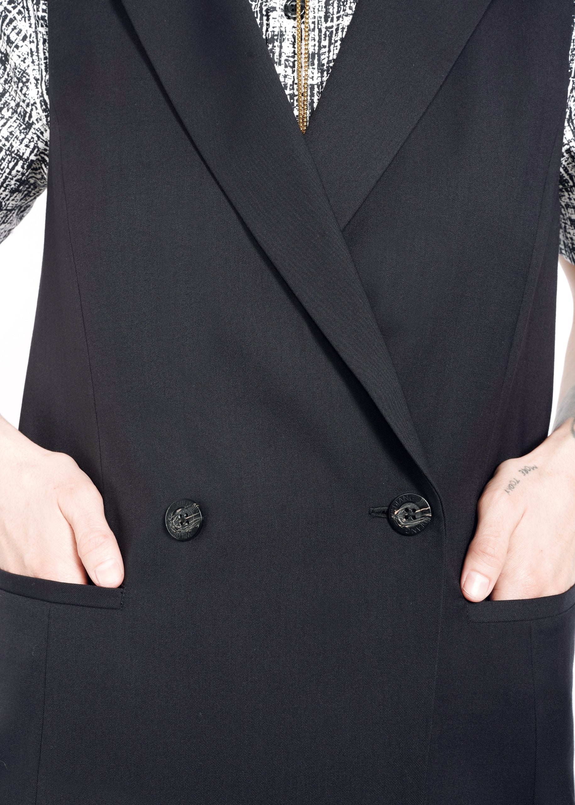 A person sporting an oversized fit with The Empower Double Breasted Vest nestles their hands in the pockets. The attire features a patterned shirt underneath, with a close-up image highlighting sleek black buttons dominating the midsection.