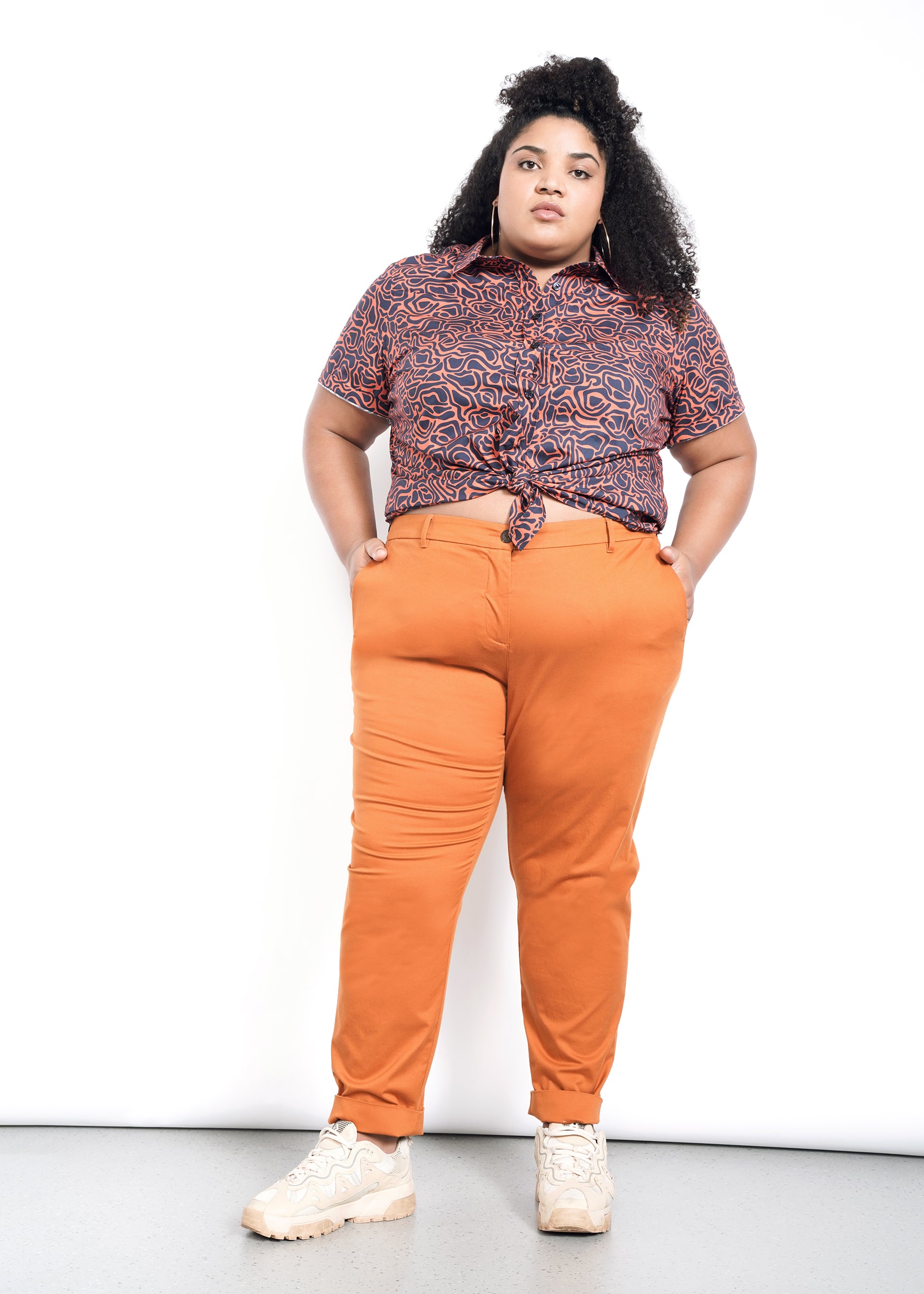 A person with curly hair confidently stands with hands in the pockets of The Essential Trouser in burnt orange, wearing a waist-tied patterned shirt with convertible cuffs, and beige sneakers, against a plain white backdrop.