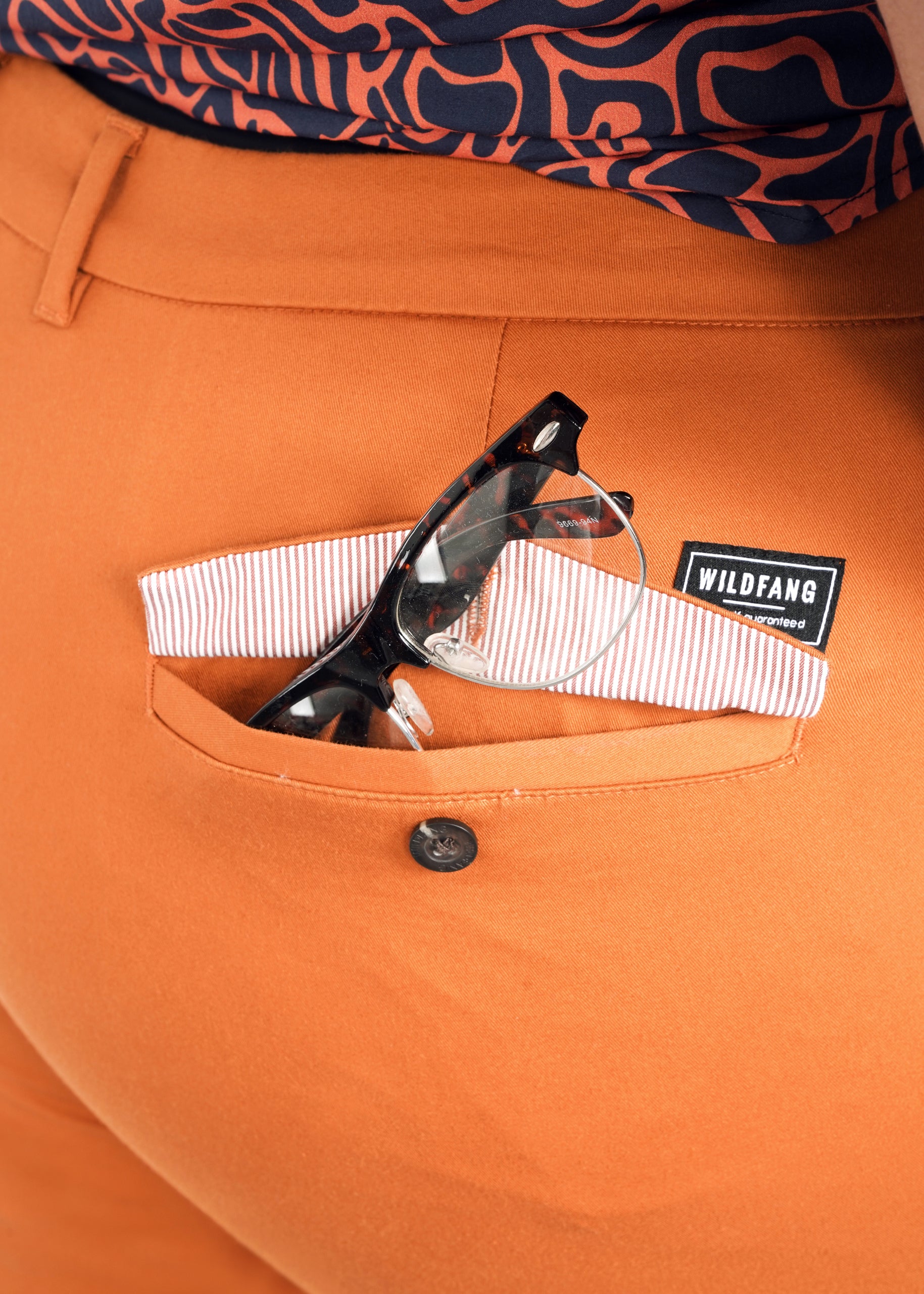 Close-up of someone in The Essential Trouser, relaxed-fit orange trousers paired with a black and white patterned shirt. Black-framed glasses and a striped cloth are partially tucked into the pocket, labeled Wildfang.