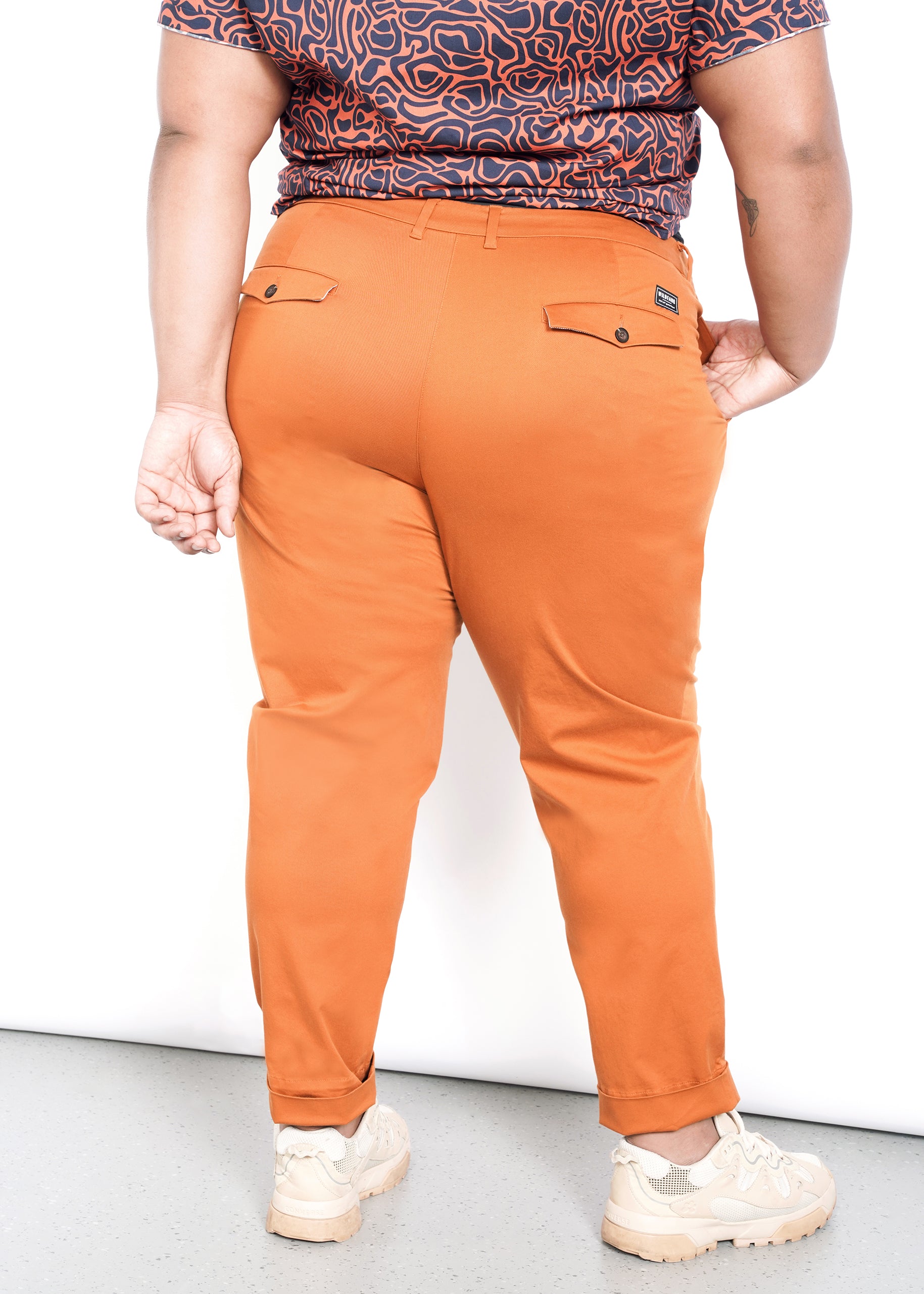 A person facing away from the camera wears The Essential Trouser in orange with a patterned shirt. Light beige sneakers finish off the look. Theyre indoors against a simple white and gray backdrop.