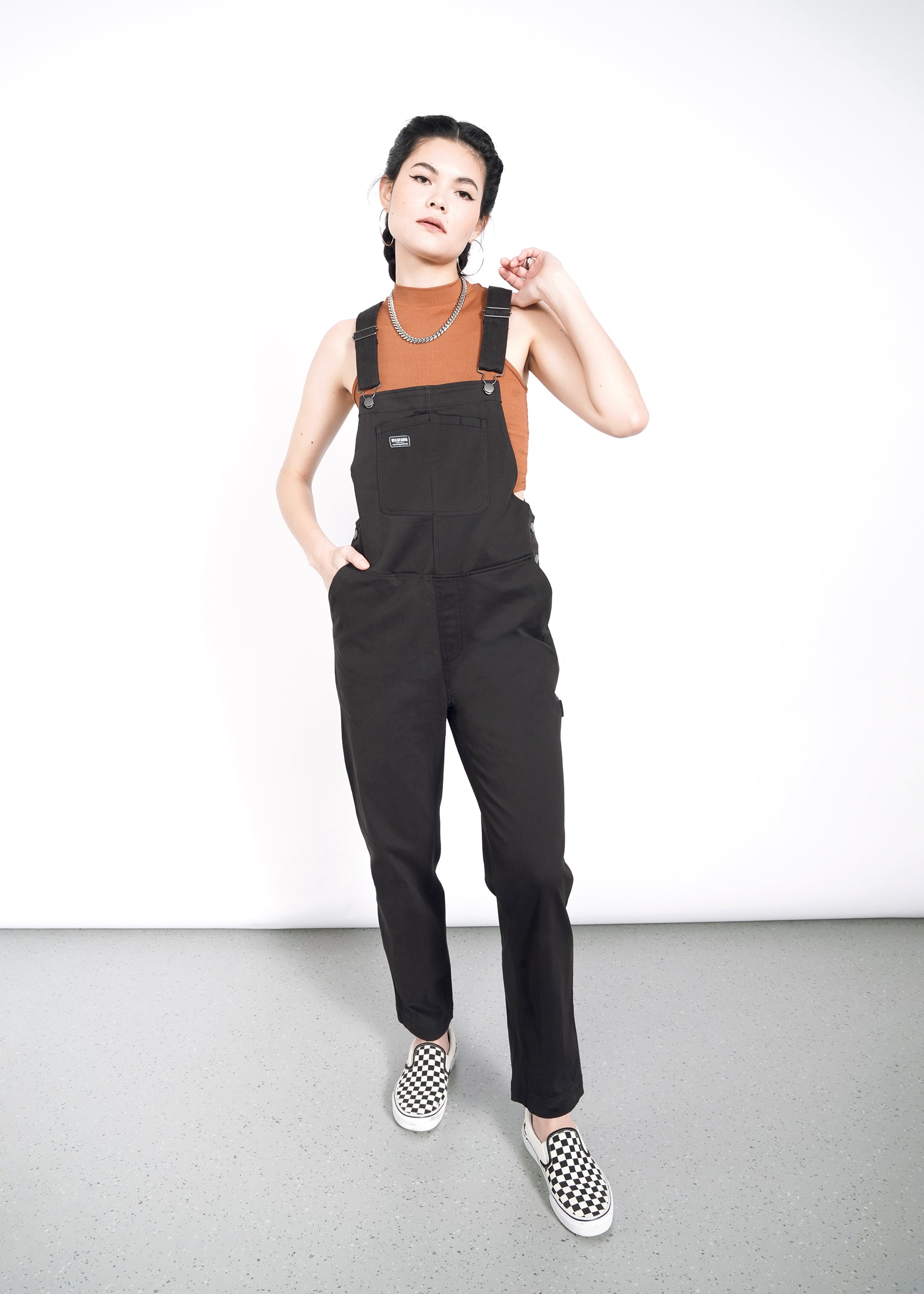 Dressed in The Essential Overall, a person stands against a white background. Their look is completed with checkered slip-on shoes and a brown sleeveless top. With braided hair, they touch their chin with one hand while the other rests in the deep pockets of their soft stretch cotton outfit.