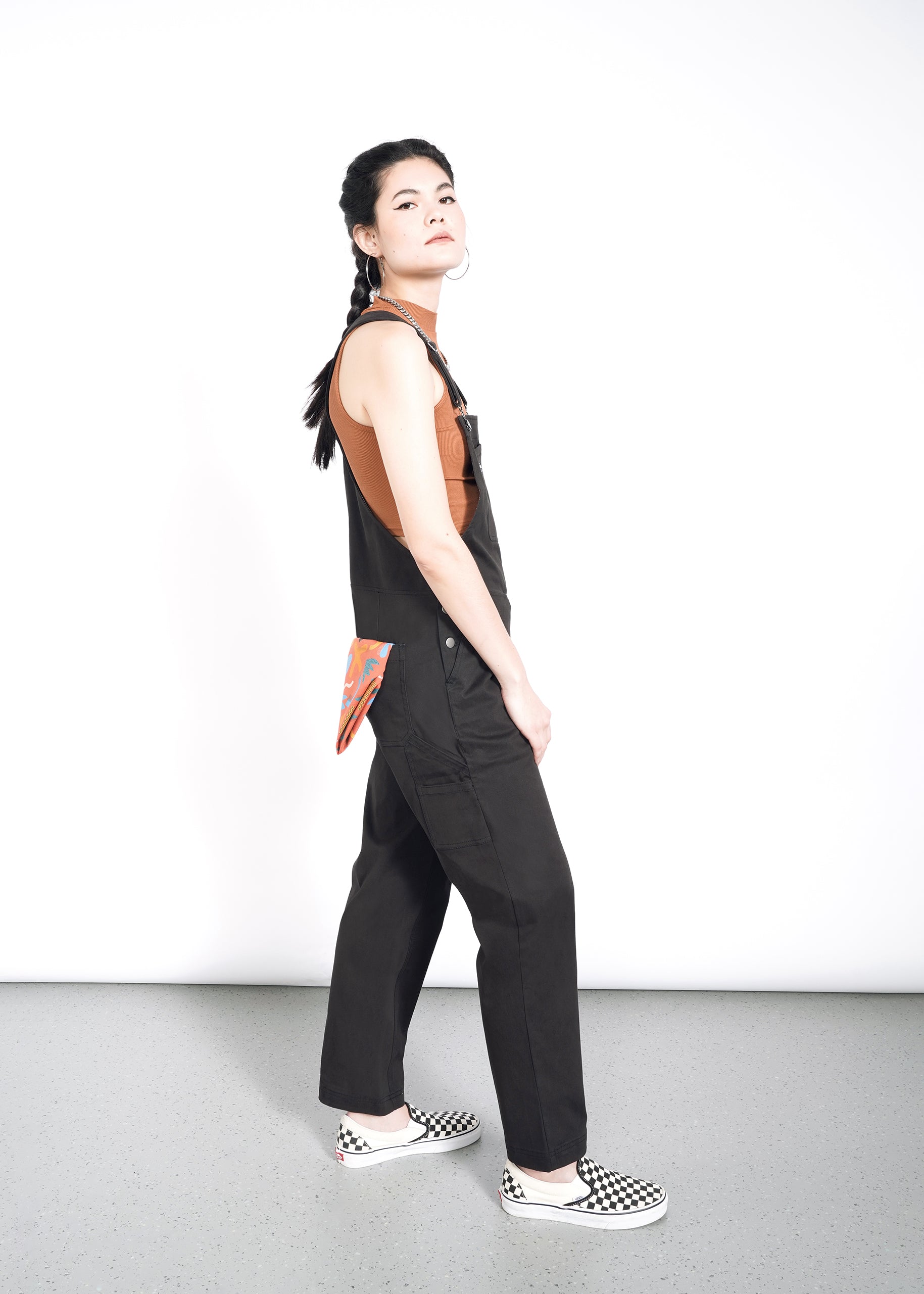 A person with braided hair stands against a white background, wearing The Essential Overall in black with deep pockets and adjustable straps over a brown tank top. A colorful handkerchief peeks from the back pocket, paired with black and white checkered slip-on shoes for a complete look.