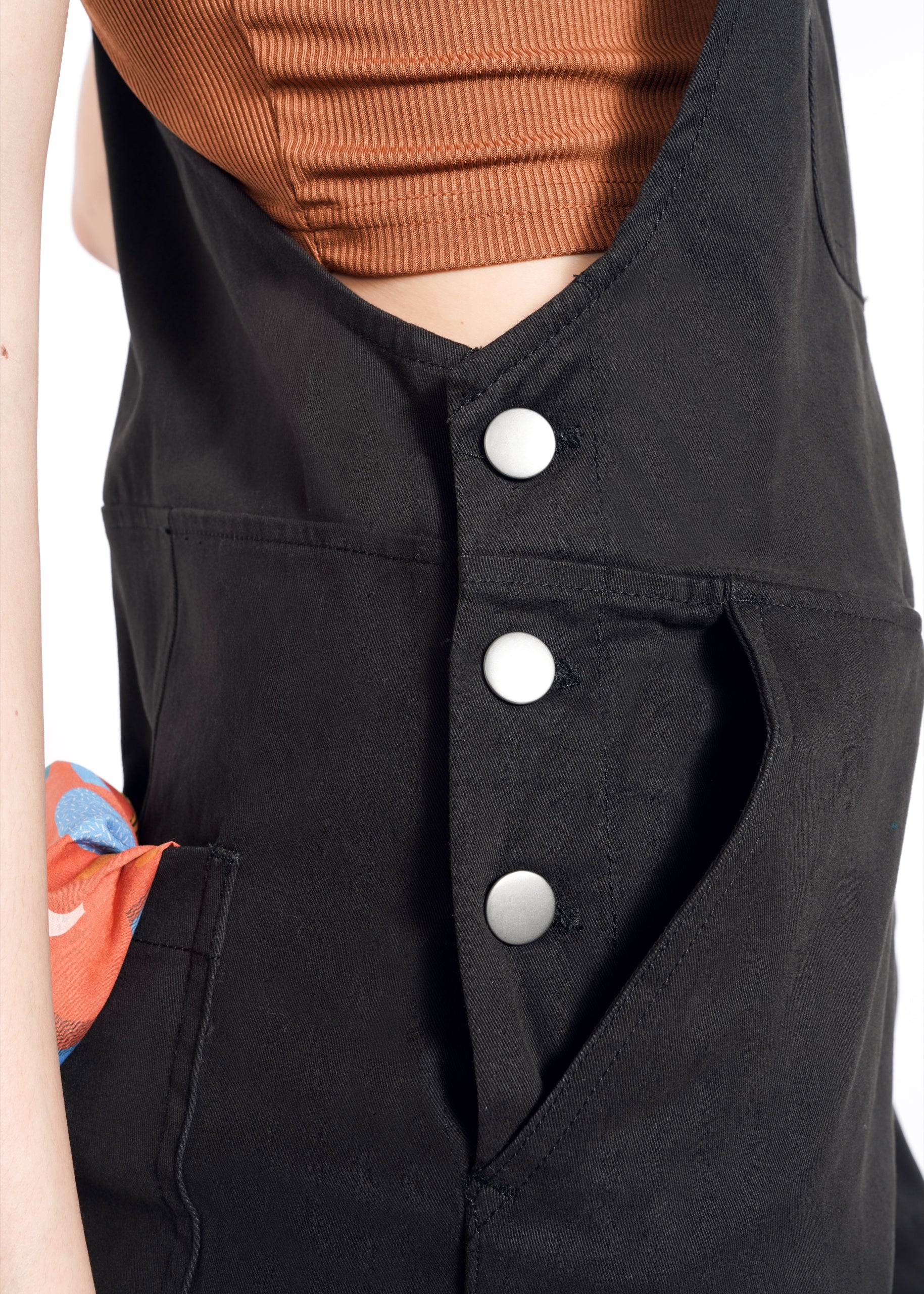 A close-up shows someone wearing The Essential Overall with adjustable straps, three silver buttons, and deep black pockets accented by a multicolored cloth. The matte-finished ribbed brown top completes the look against a plain white background.