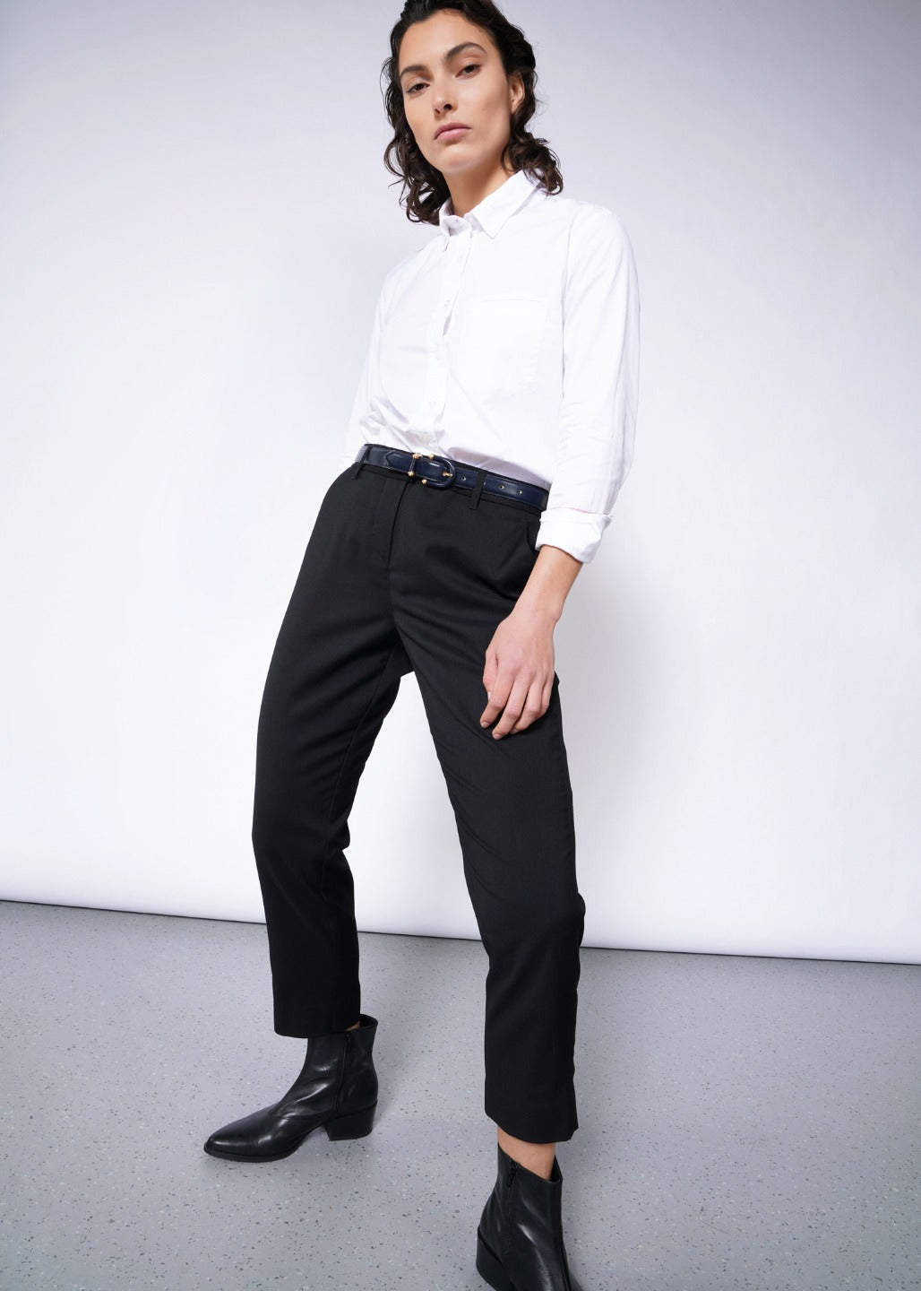 A person with short, dark wavy hair confidently poses against a white backdrop, hands on thighs. Theyre wearing The Empower Slim Crop Pant in a high-rise fit and a white shirt, paired with chic black ankle boots for an elegant look.