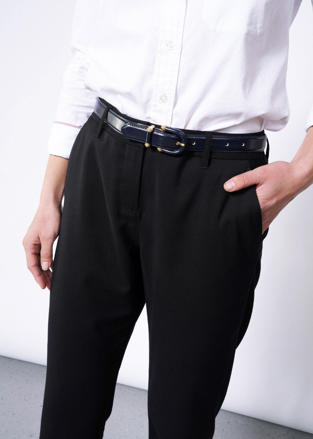 A person wearing a white button-up shirt tucked into The Empower Slim Crop Pant, secured with a black belt. They stand against a light background, one hand in pocket, effortlessly exuding professional style.