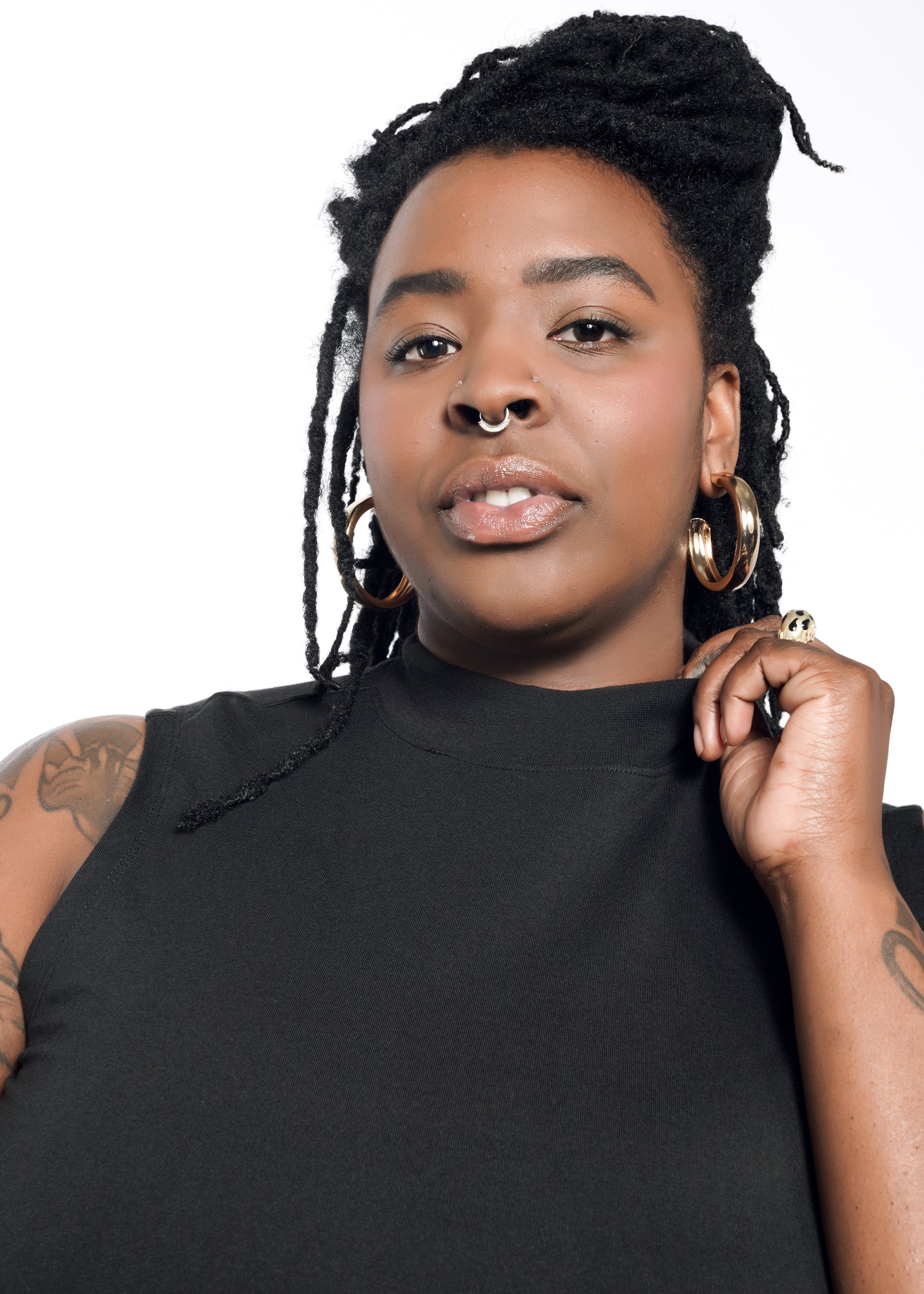 A person with long braided hair and a septum piercing confidently faces the camera. They wear The Essential Mock Neck Tank, a sleeveless black cotton piece, along with gold hoop earrings and visible arm tattoos. A plain white backdrop highlights this relaxed-fit tank as an ideal layering piece.