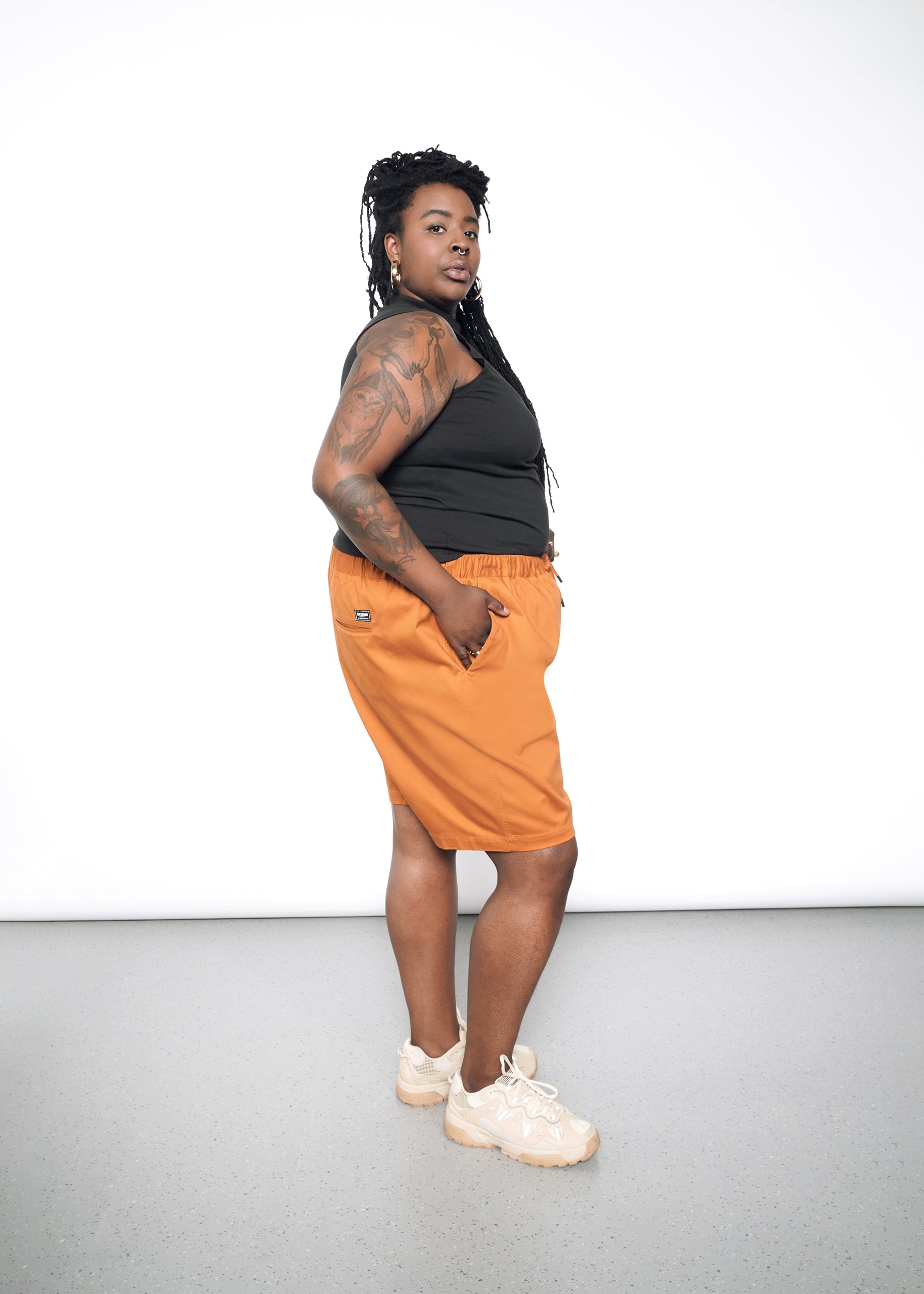 A person with long hair and arm tattoos wears The Essential Mock Neck Tank in relaxed-fit black cotton, paired with orange shorts and sneakers, stands sideways on a plain gray floor and looks at the camera against a white background.