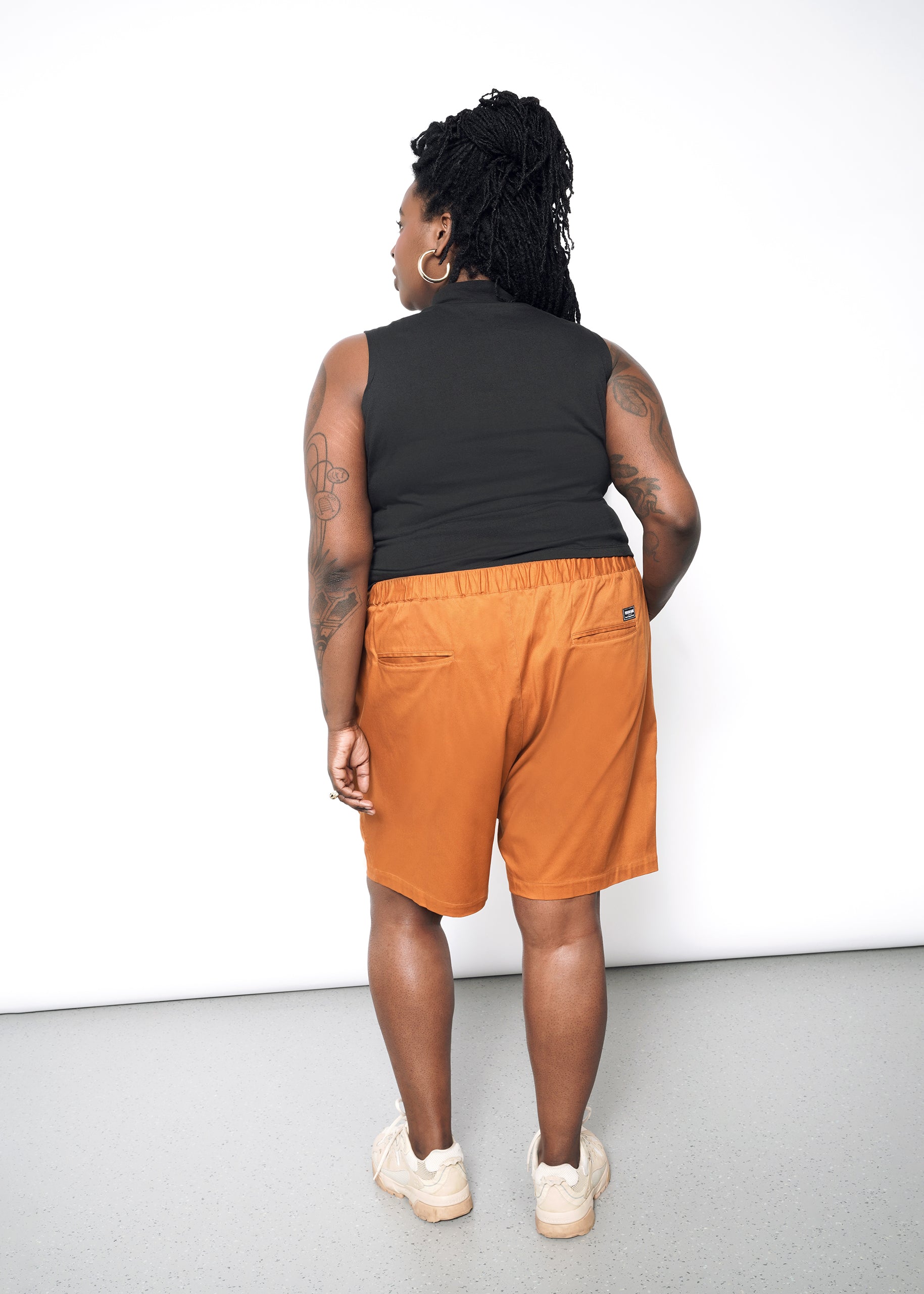 A person with braided hair, facing away, wears The Essential Mock Neck Tank, along with orange shorts and beige sneakers. Their tattoos are visible as they stand against a plain white wall.