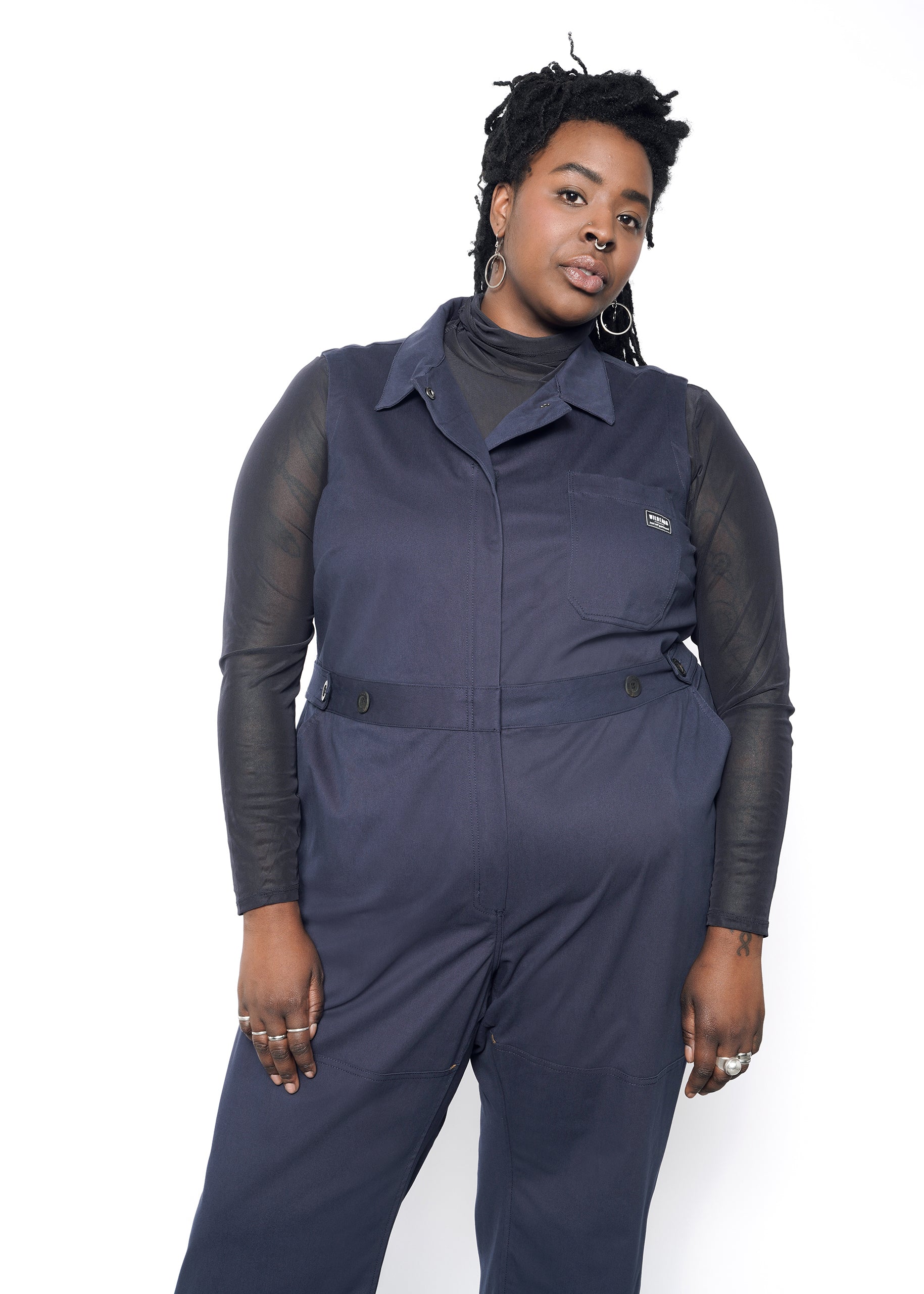 A person wears The Essential Sleeveless High Waisted Coverall in navy blue with a cotton stretch fabric over a transparent black long-sleeve top. They pose against a white backdrop with long braided hair, hoop earrings, and multiple rings.