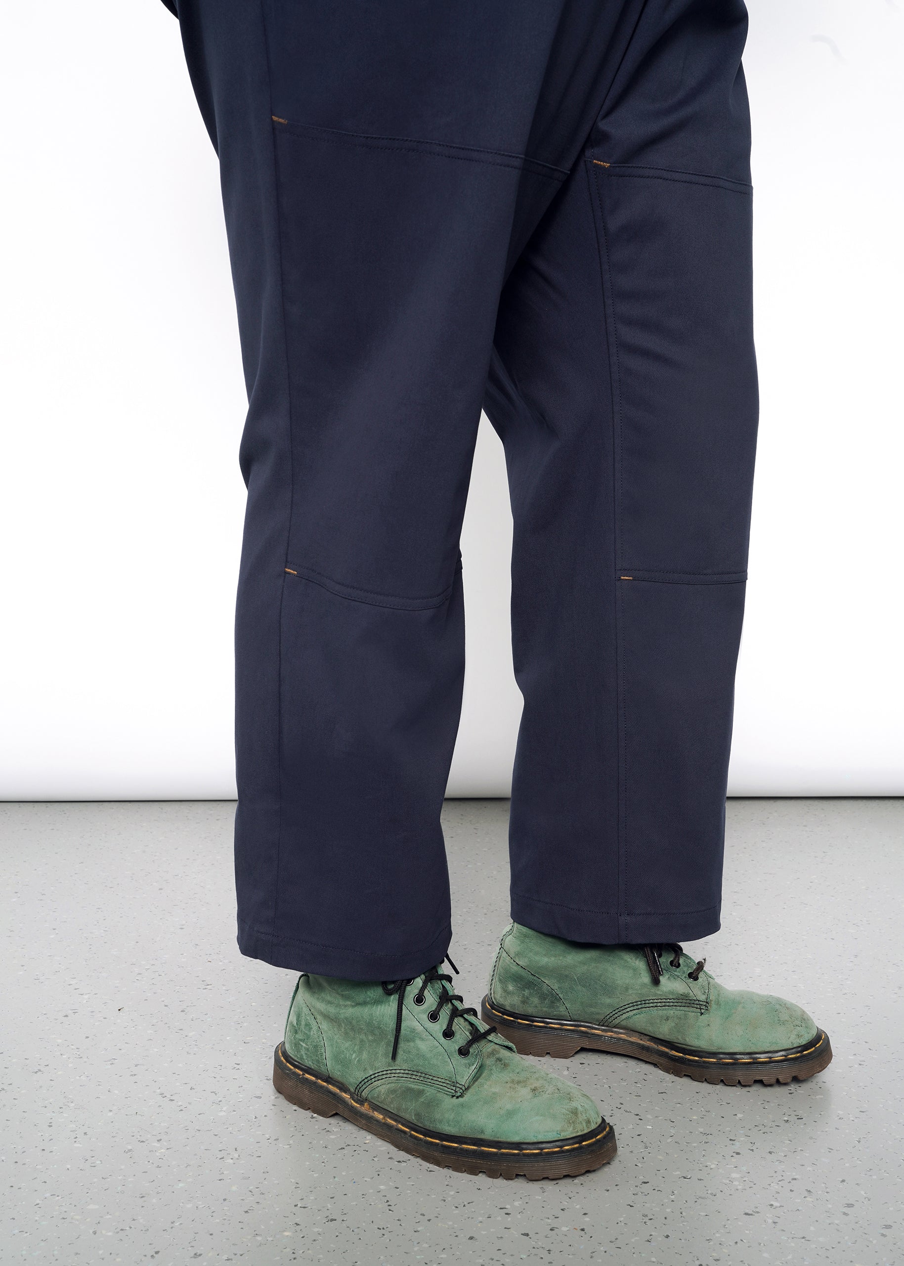 A person wears The Essential Sleeveless High Waisted Coverall in dark blue with green lace-up suede boots, standing on a speckled gray floor. The white background accentuates the contrast between the cotton stretch coverall and the shoes.