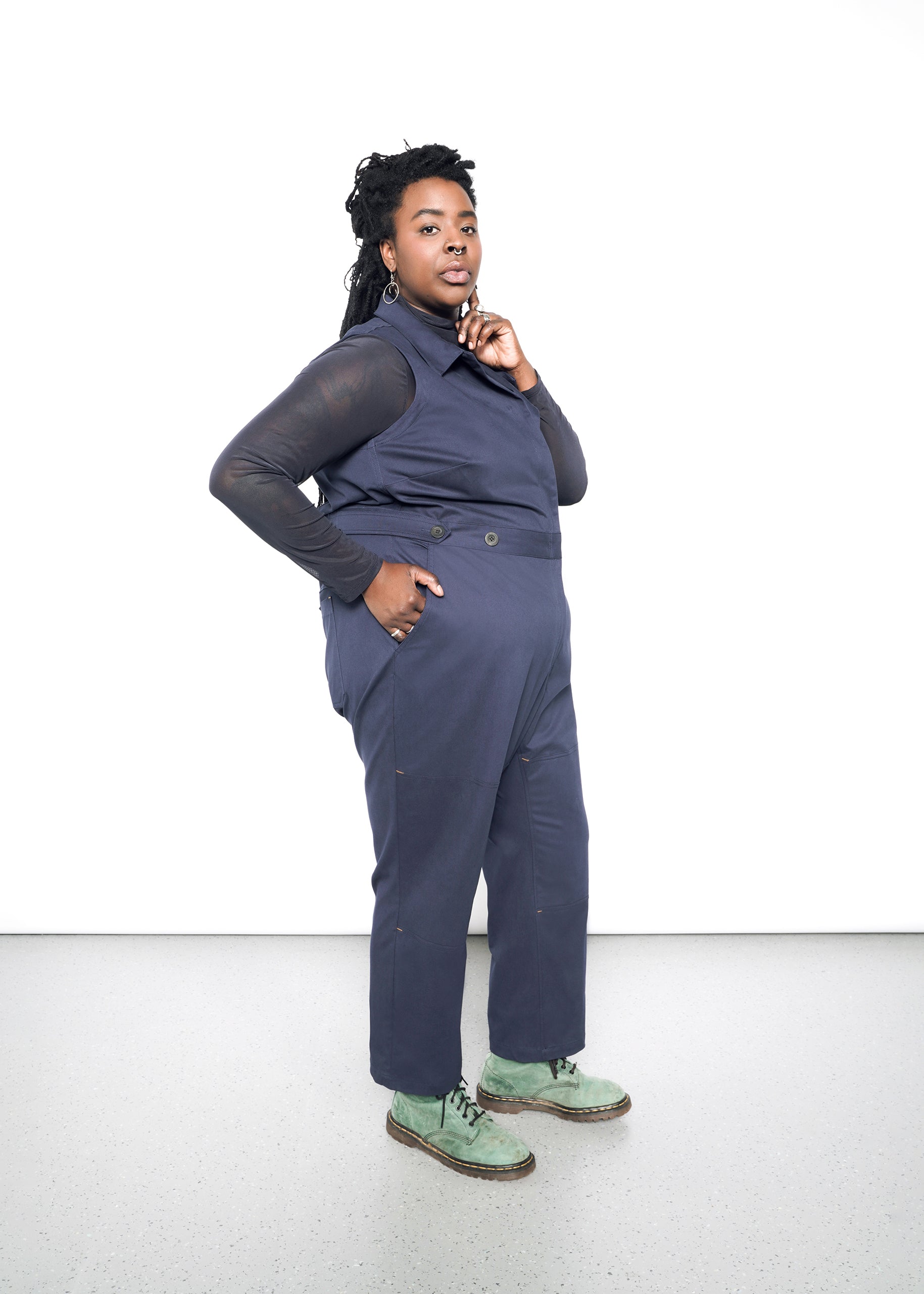 A person confidently poses against a white background, wearing The Essential Sleeveless High Waisted Coverall in dark blue over a long-sleeve shirt and green shoes. With shoulder-length dark braided hair, they rest their chin on their hand.