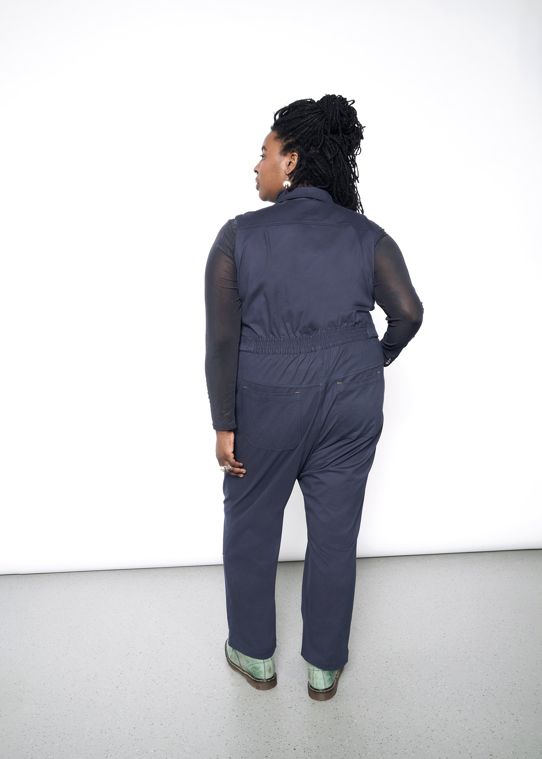 A person with braided hair stands facing away, wearing The Essential Sleeveless High Waisted Coverall in dark navy and light green shoes. The high-waisted design complements the plain white background, while the floor remains gray.