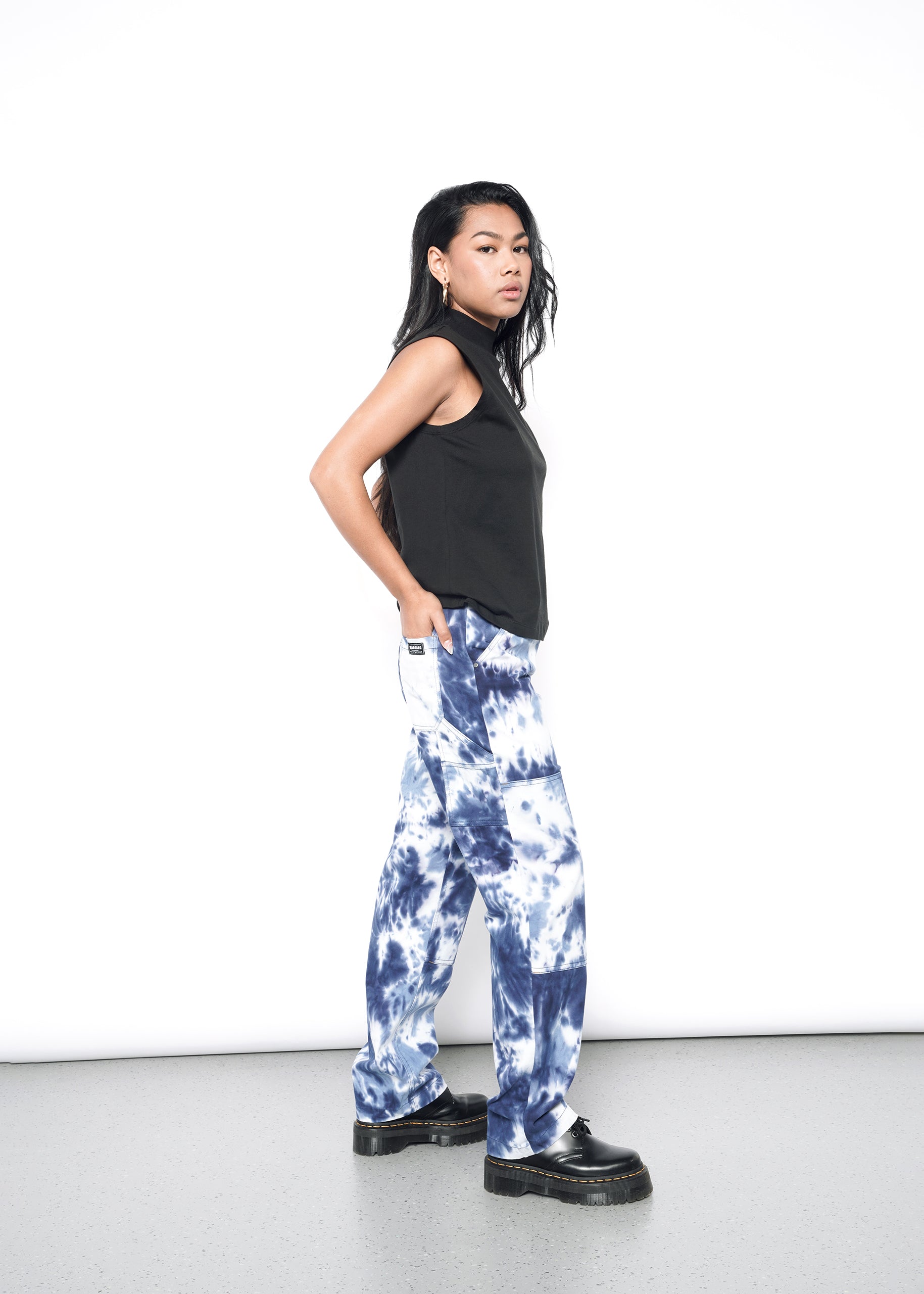 A person with long dark hair stands confidently against a white background, wearing The Essential Mock Neck Tank for layering, tie-dye blue and white cotton pants, and black platform shoes, hands in pockets.