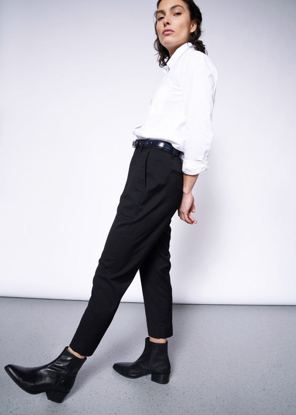 Person in a light gray setting, wearing a white shirt and The Empower Slim Crop Pant in black with a high-rise fit, paired with black ankle boots. They gaze confidently forward, slightly leaning back.