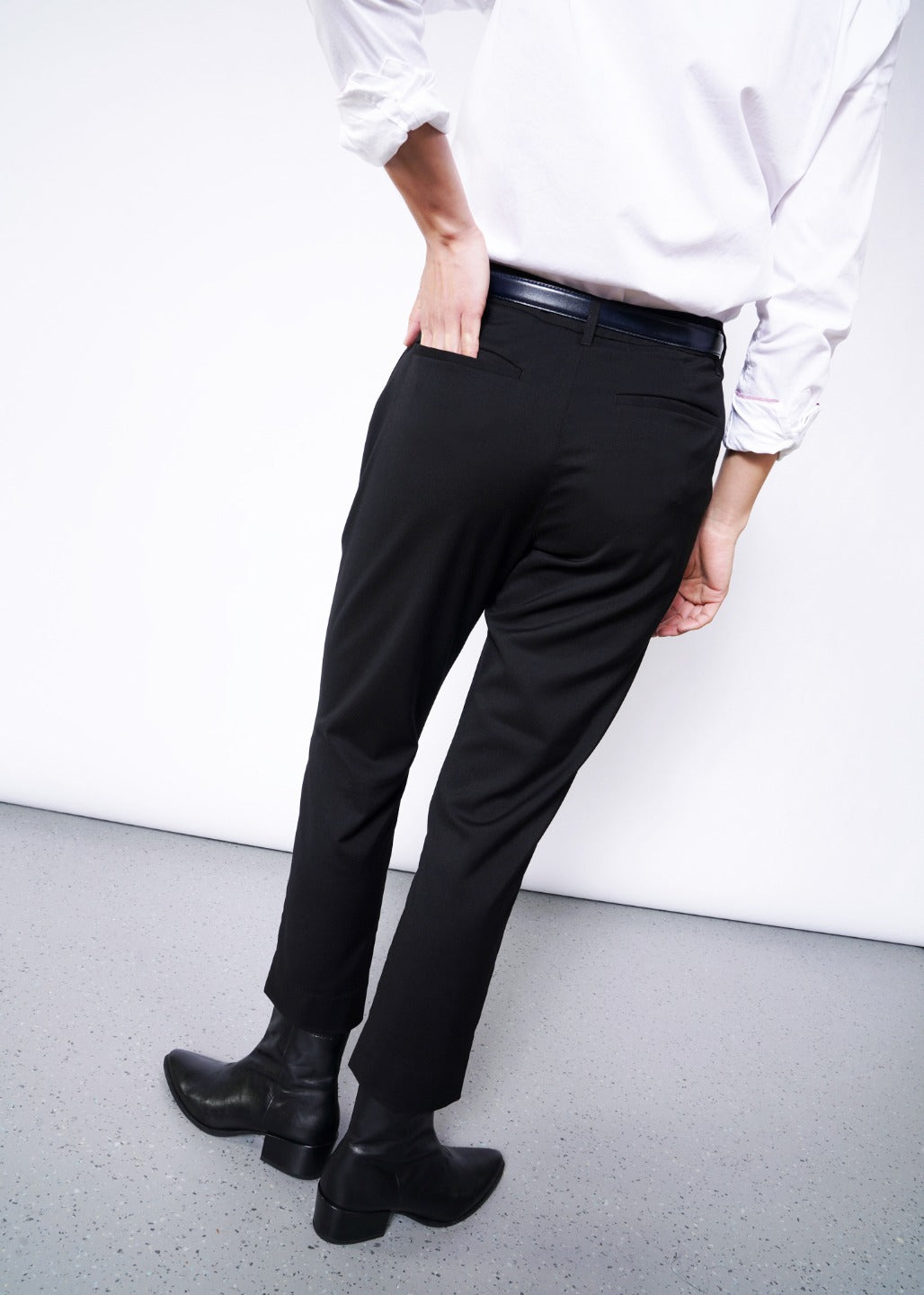 A person wearings The Empower Slim Crop Pant with a white shirt and black ankle boots stands against a plain white wall on a gray floor, exuding stylish simplicity.