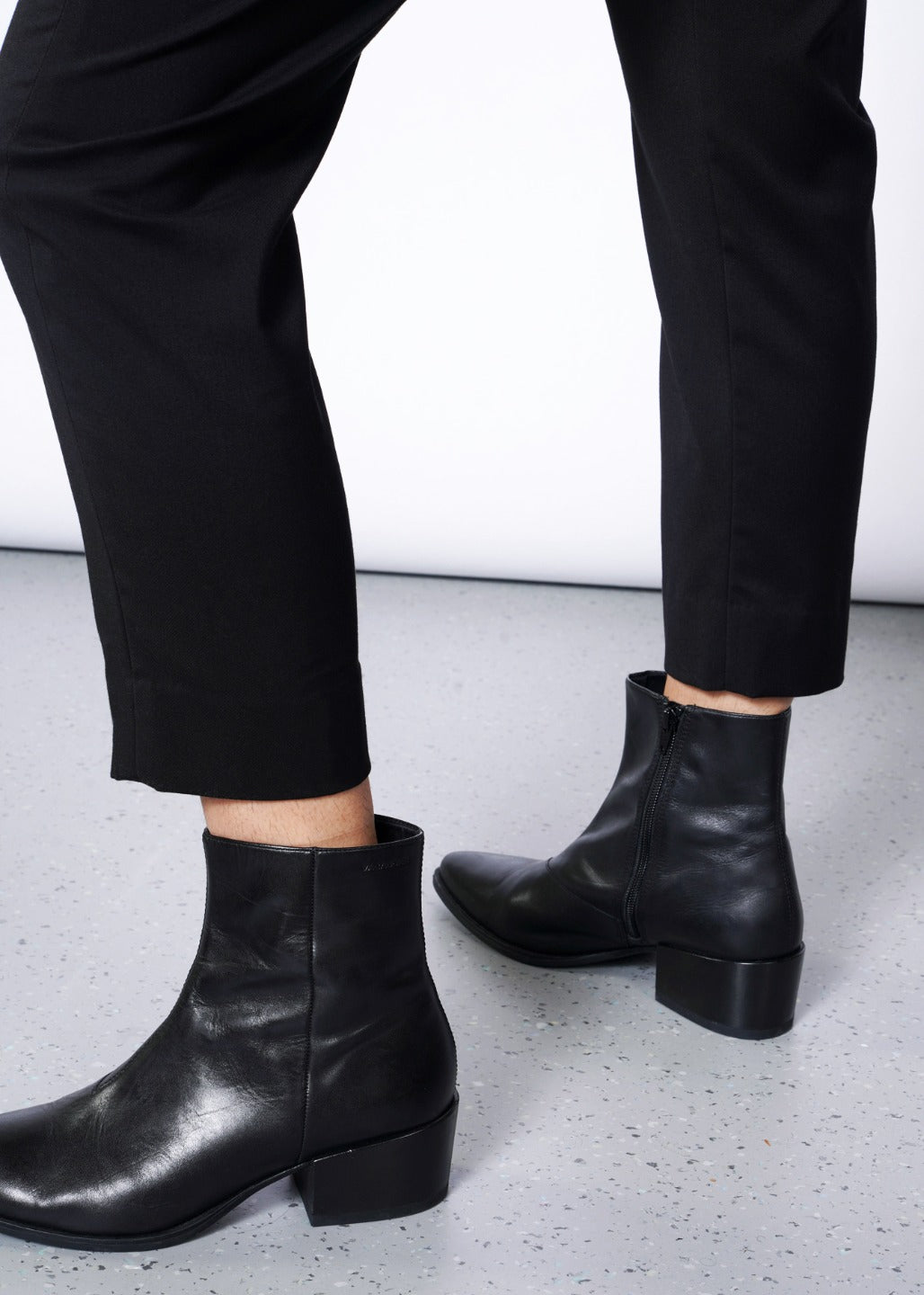 Wearing The Empower Slim Crop Pant in sleek black and black leather ankle boots with side zippers, a person stands confidently on a light gray speckled floor.