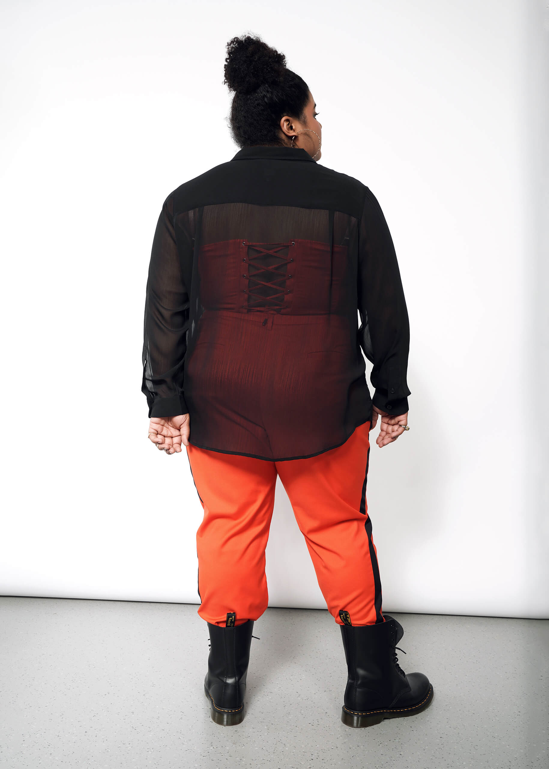 Against a white backdrop, a person is seen from behind wearing The Empower Sheer Long Sleeve Button Up in black. Paired with bright orange pants tucked into black boots and hair in a bun, the corset-style back adds an edgy yet chic vibe.