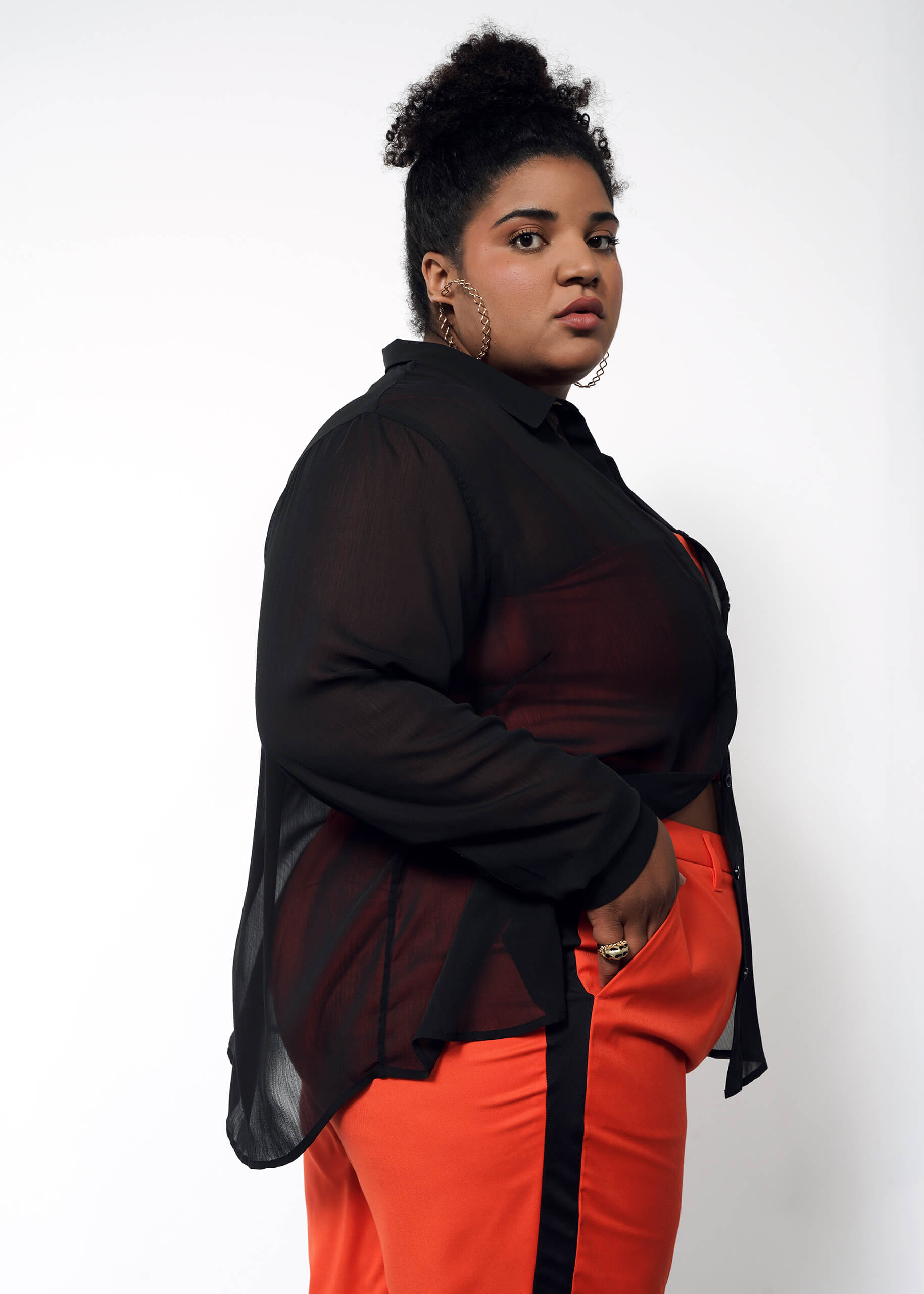 With curly hair, they confidently wear The Empower Sheer Long Sleeve Button Up, an oversized sheer black blouse over a black top, paired with high-waisted orange pants. Their hoop earrings and rings complete the look against a plain white background.