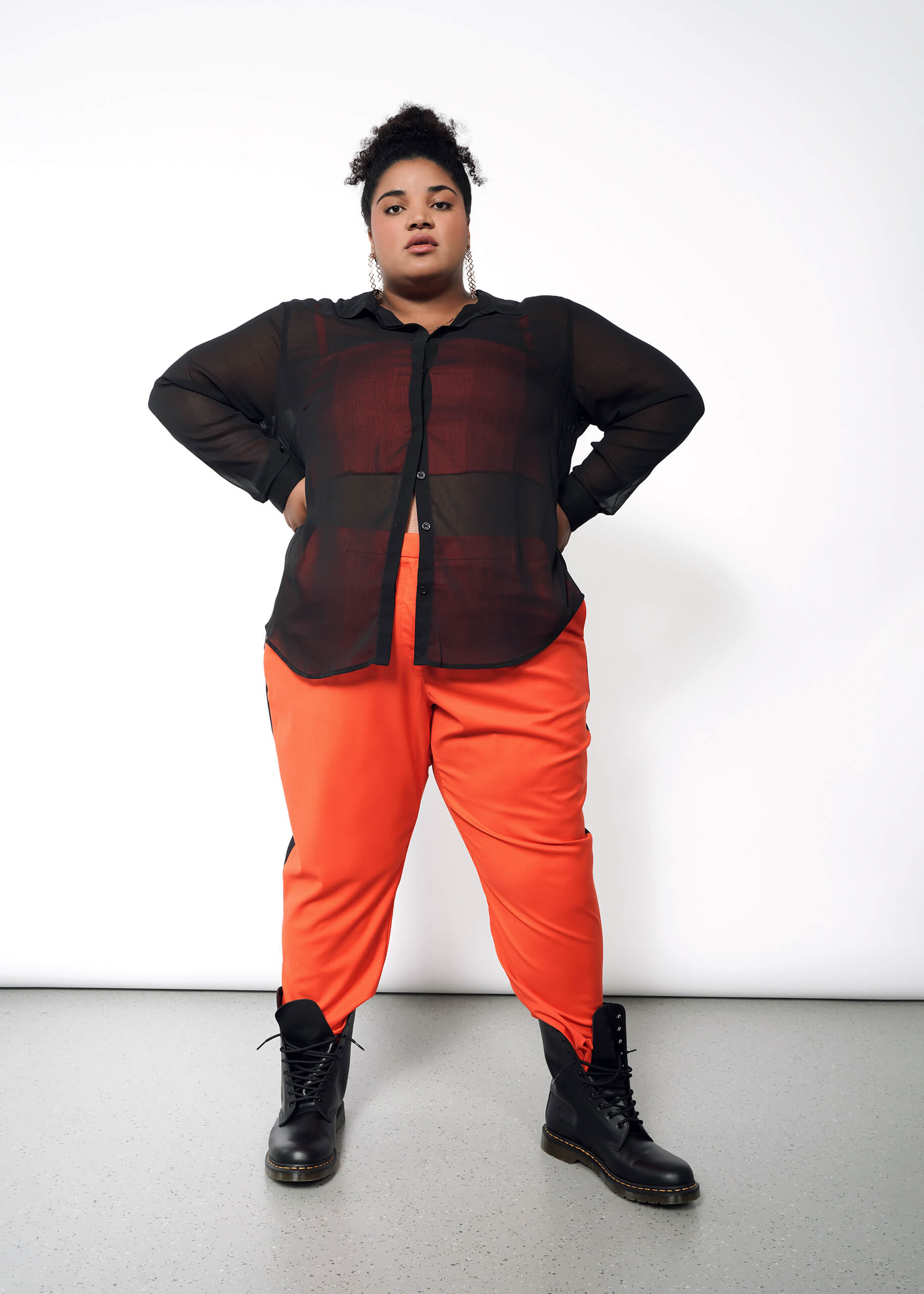 Wearing The Empower Sheer Long Sleeve Button Up with an oversized fit over a button-closure shirt, bright orange pants, and black boots, the person stands confidently against a white background. Their curly hair is styled up with large earrings as accessories.