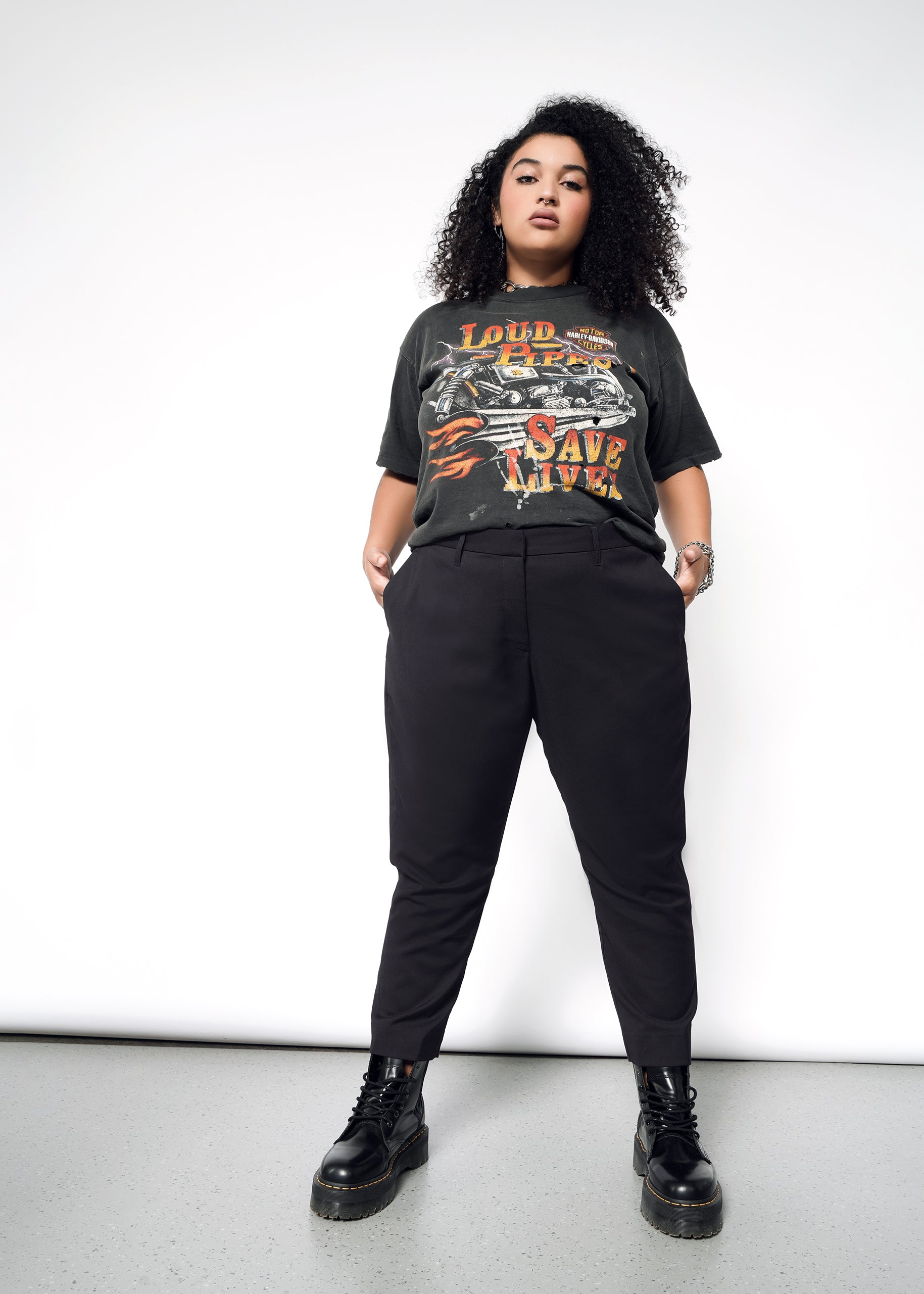 A person with curly hair confidently stands with hands in pockets, wearing a graphic T-shirt, The Empower Slim Crop Pant, and black boots. The high-rise fit accentuates their style against the plain white background.