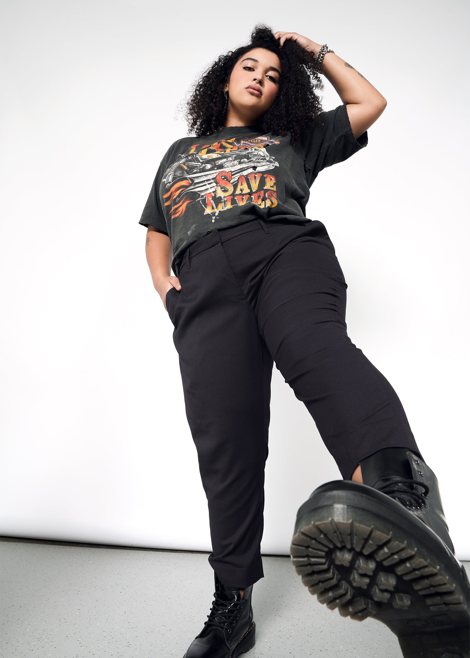 A curly-haired woman poses confidently in a graphic tee, The Empower Slim Crop Pant with a high-rise fit, and black boots. With one hand in her pocket and a foot extended forward, she stands against a plain background.