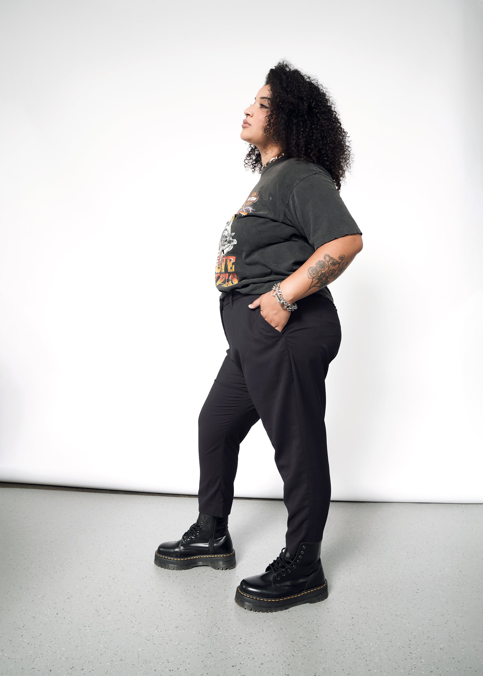 A person with curly hair in profile against a plain backdrop wears a black graphic T-shirt, The Empower Slim Crop Pant in black, and black platform boots. Tattoos cover their left arm while they gaze upward with one hand in their pocket.