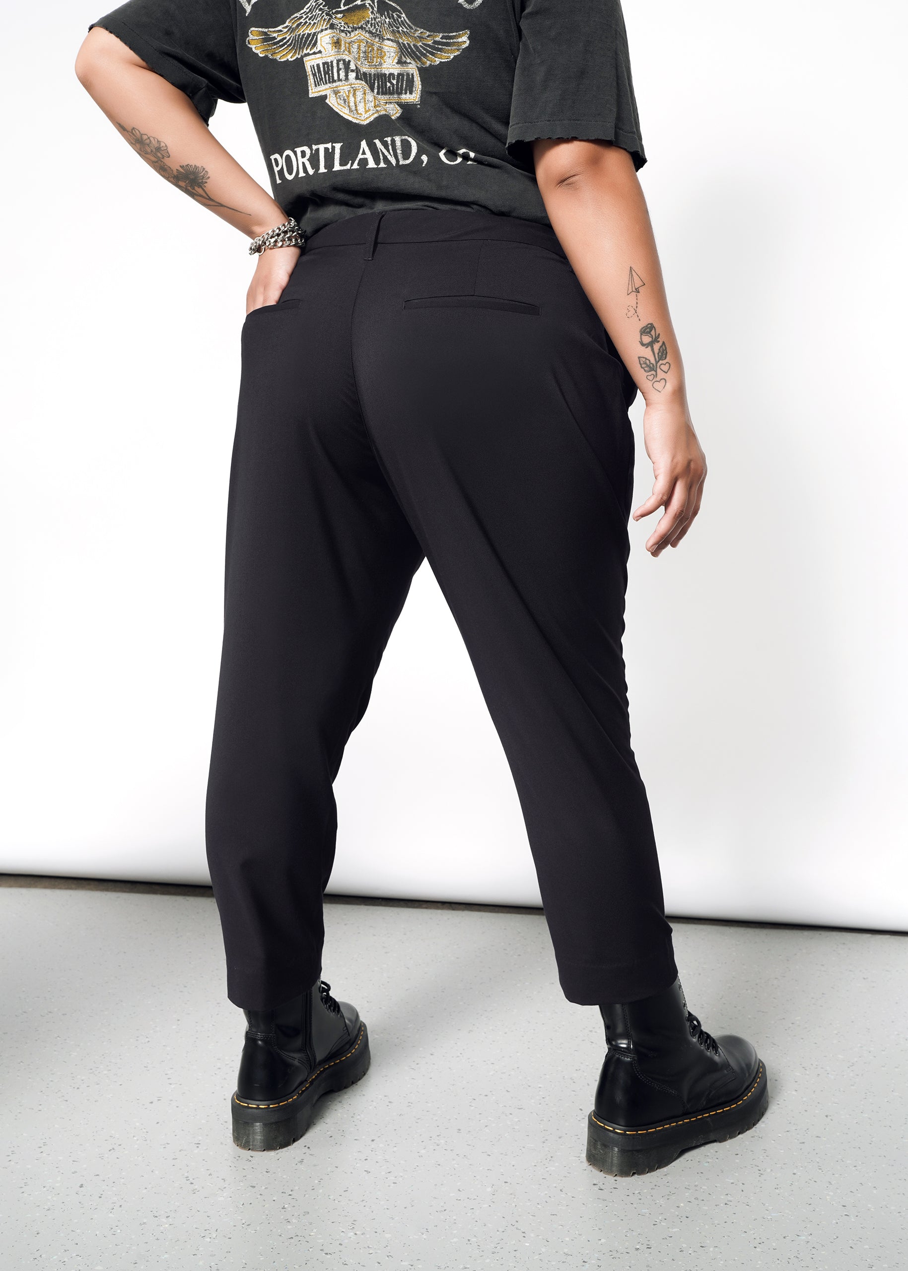 Facing away from the camera against a light-colored background, a person showcases The Empower Slim Crop Pant in black, paired with black boots and a black T-shirt adorned with a design and text. Their tattooed arms are visible as the slim crop pants end stylishly at ankle length.