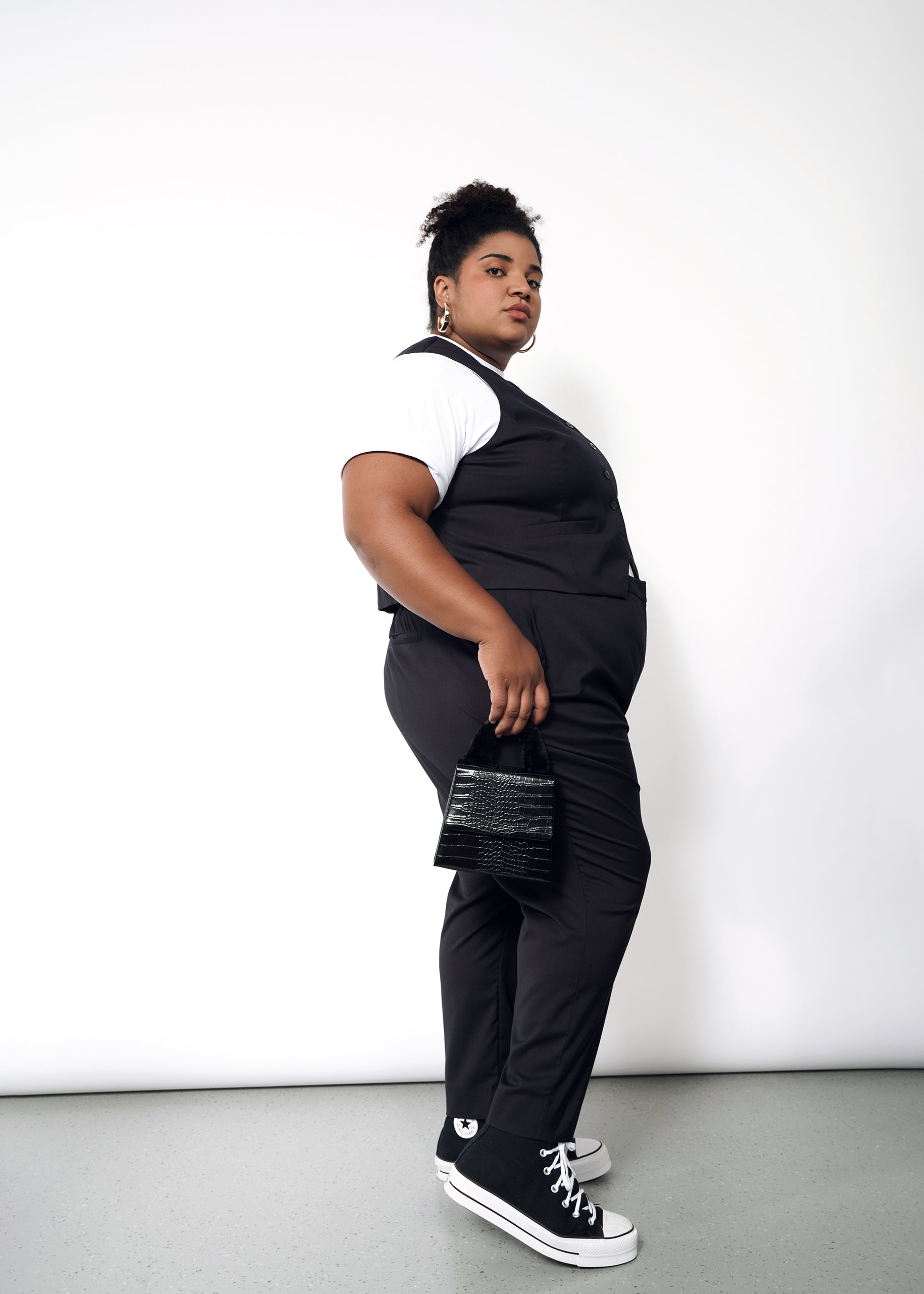 Against a white wall, a person confidently showcases The Empower Vest with full stretch lining and tailored fit over a white t-shirt. The look is completed with trousers, sneakers, a small black bag, and an updo hairstyle, exuding stylishness and poise.