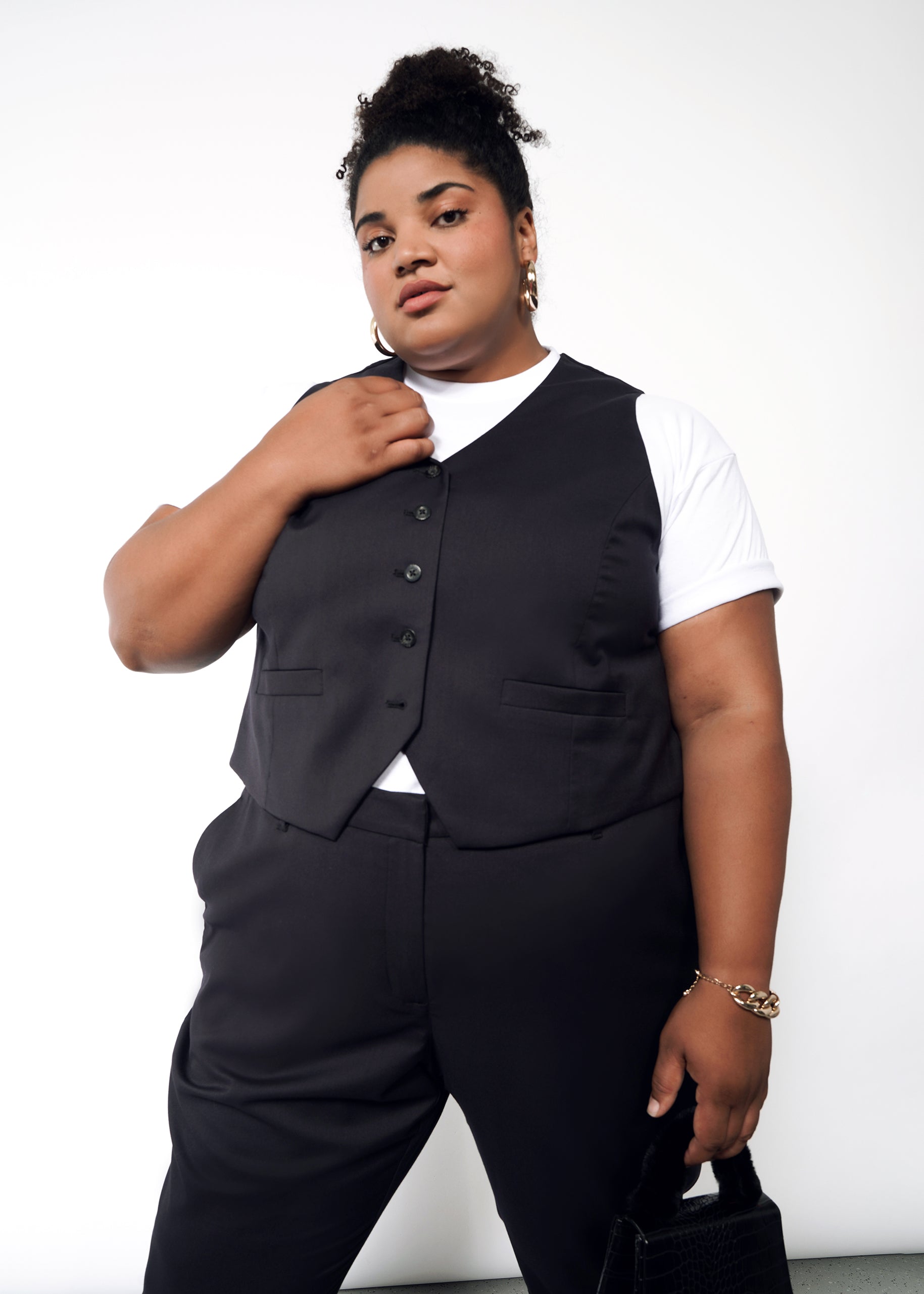 With confidence, a curly-haired individual wears The Empower Vest over a white shirt and black pants. They carry a black handbag, wear a gold bracelet, and pose against a plain white backdrop.